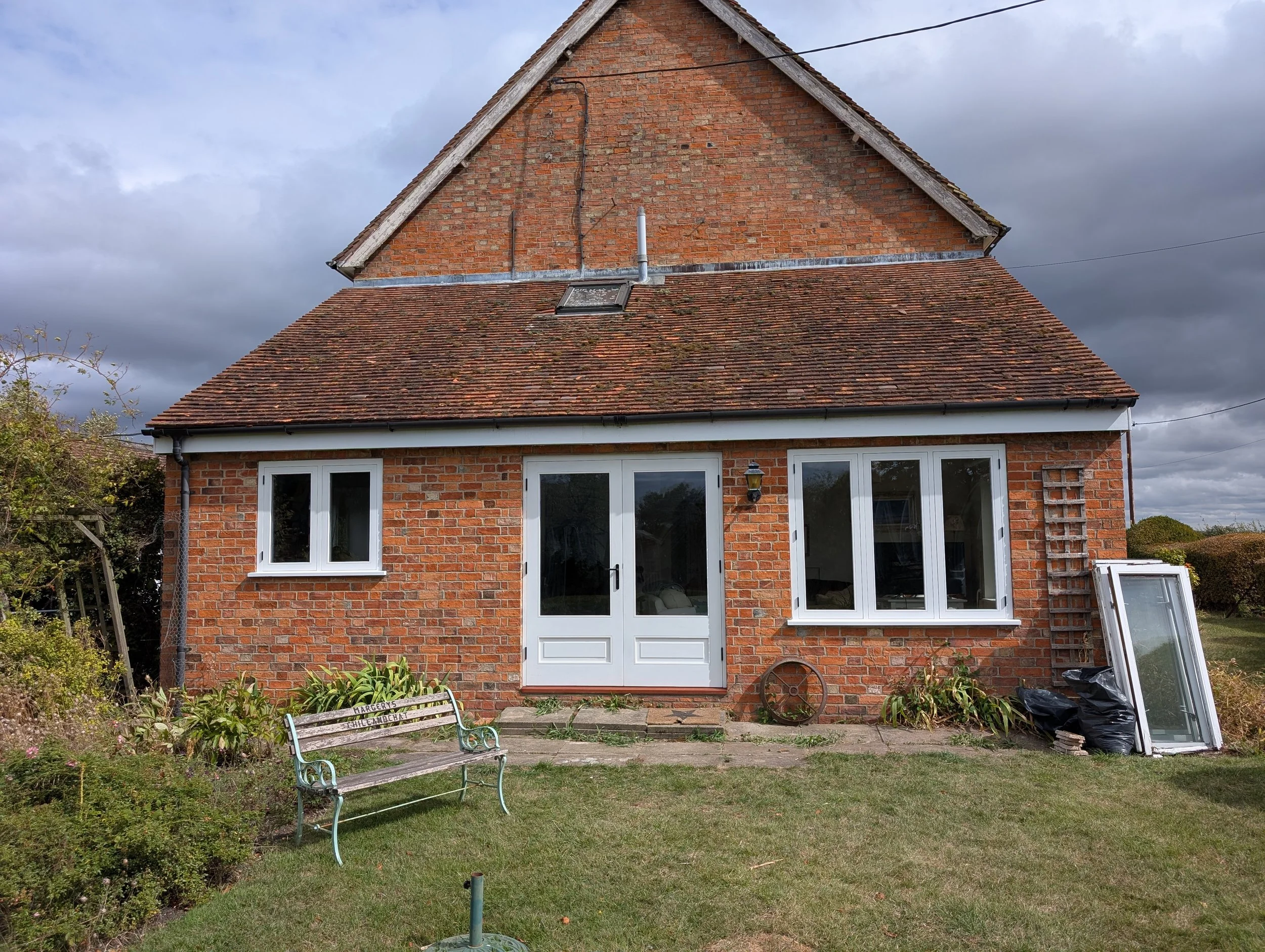 Brick house with white door and windows, garden with a bench, and a cloudy sky.