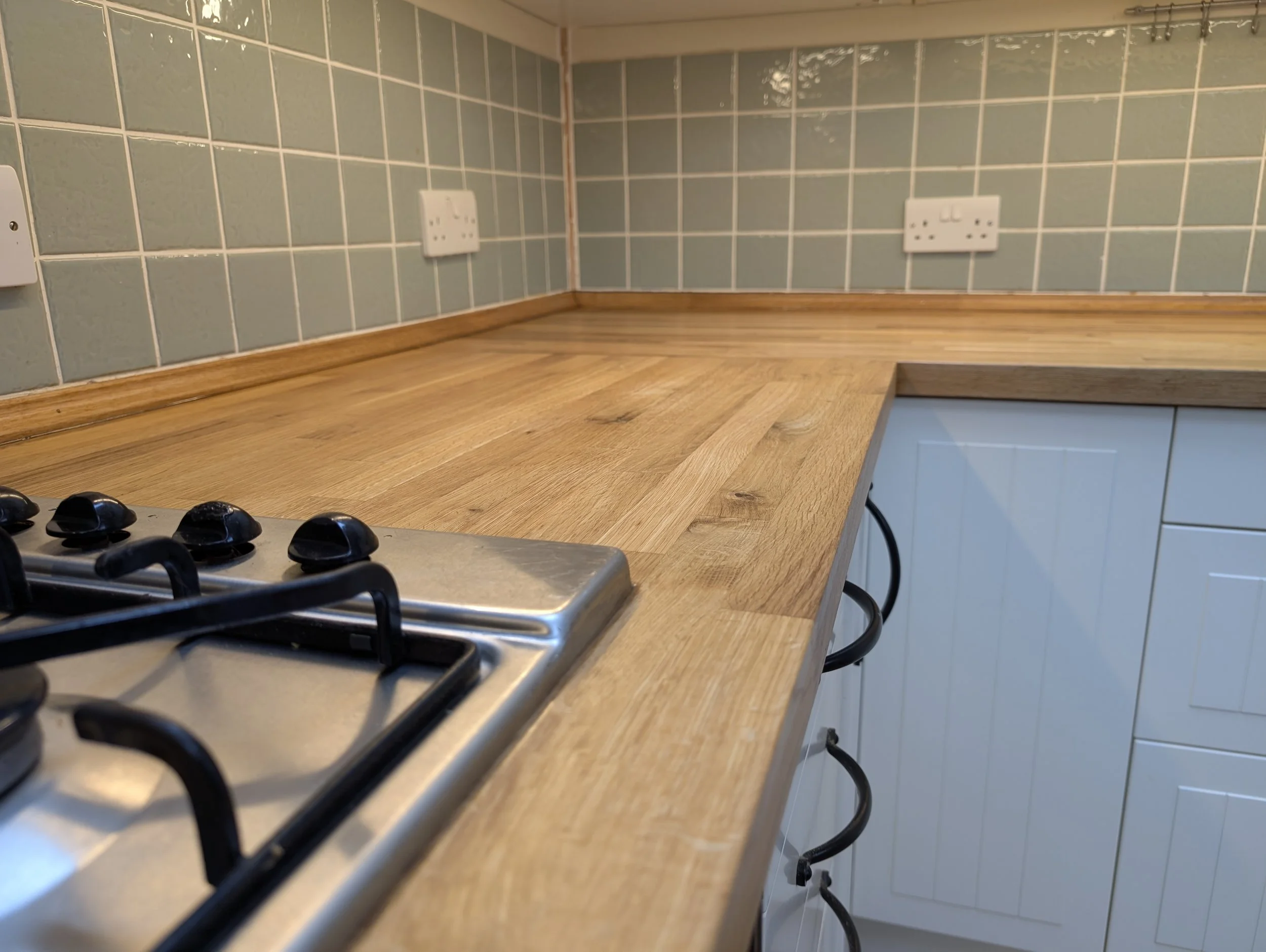 Close-up of wooden kitchen countertop with a stainless steel gas stove in the foreground and light green tiled backsplash with electrical outlets in the background.