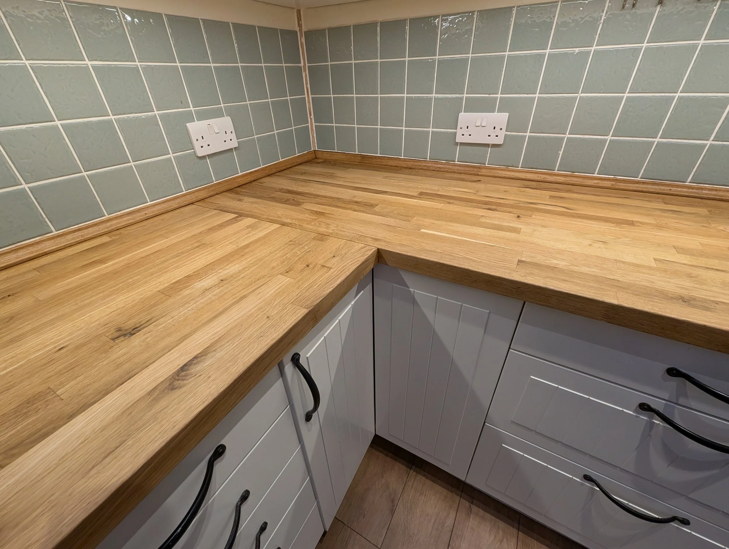 Kitchen countertop with wooden surface, white cabinets, grey tiled backsplash, and electrical outlets on the wall.