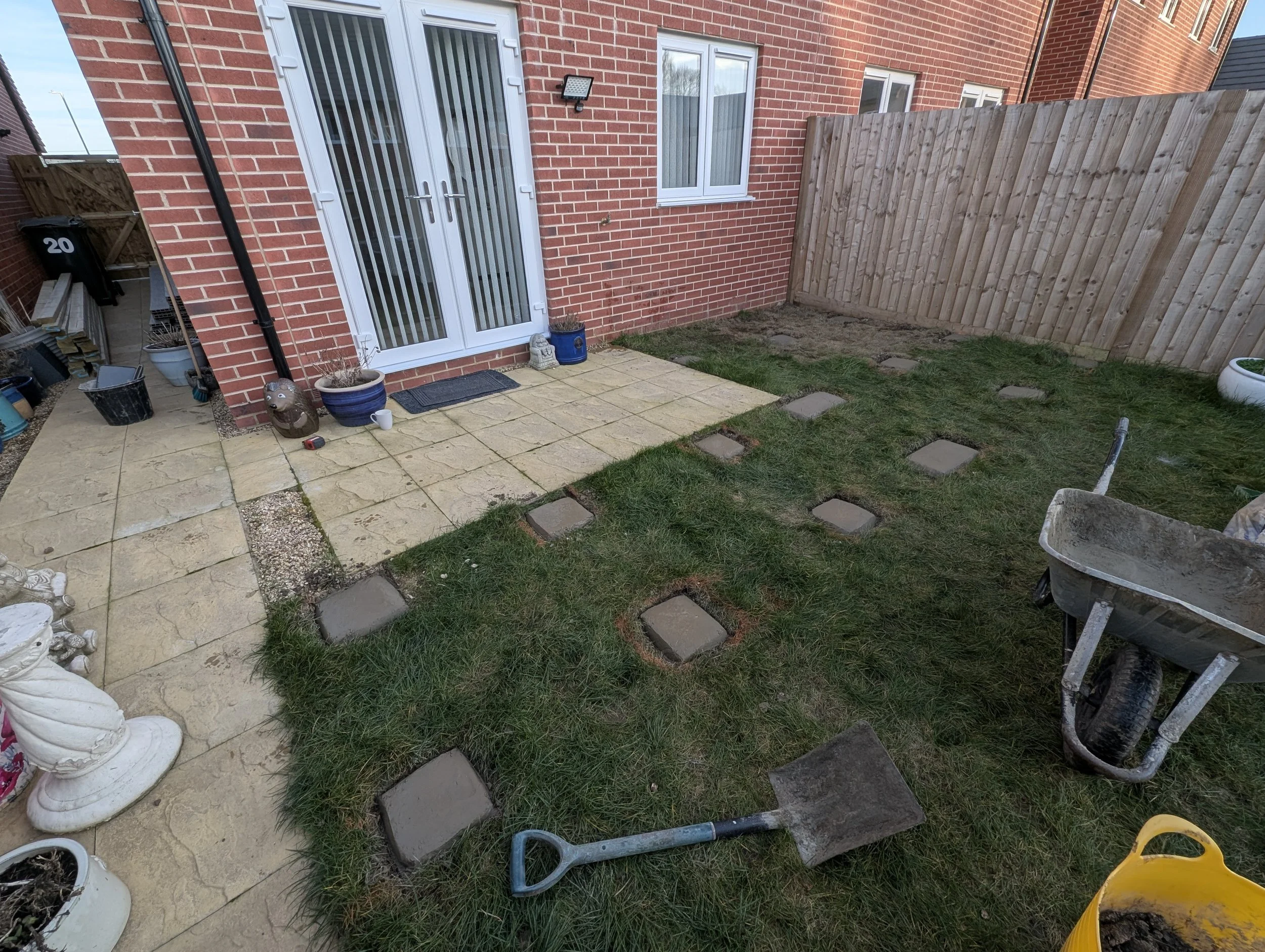Backyard with a partially paved area, a grassy patch with stepping stones, a wheelbarrow, planting tools, and potted plants next to a brick house with a wooden fence.