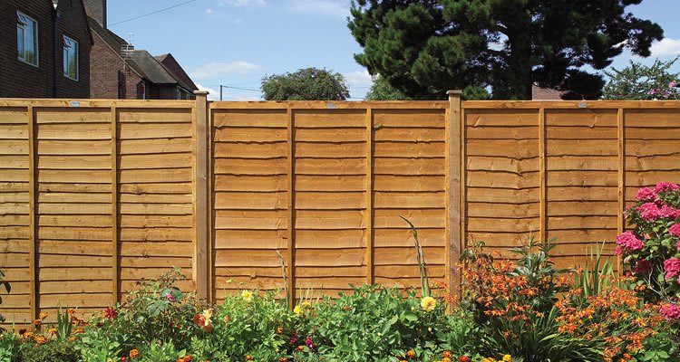 Wooden privacy fence with flowering plants and garden in front, neighborhood houses and trees in the background.