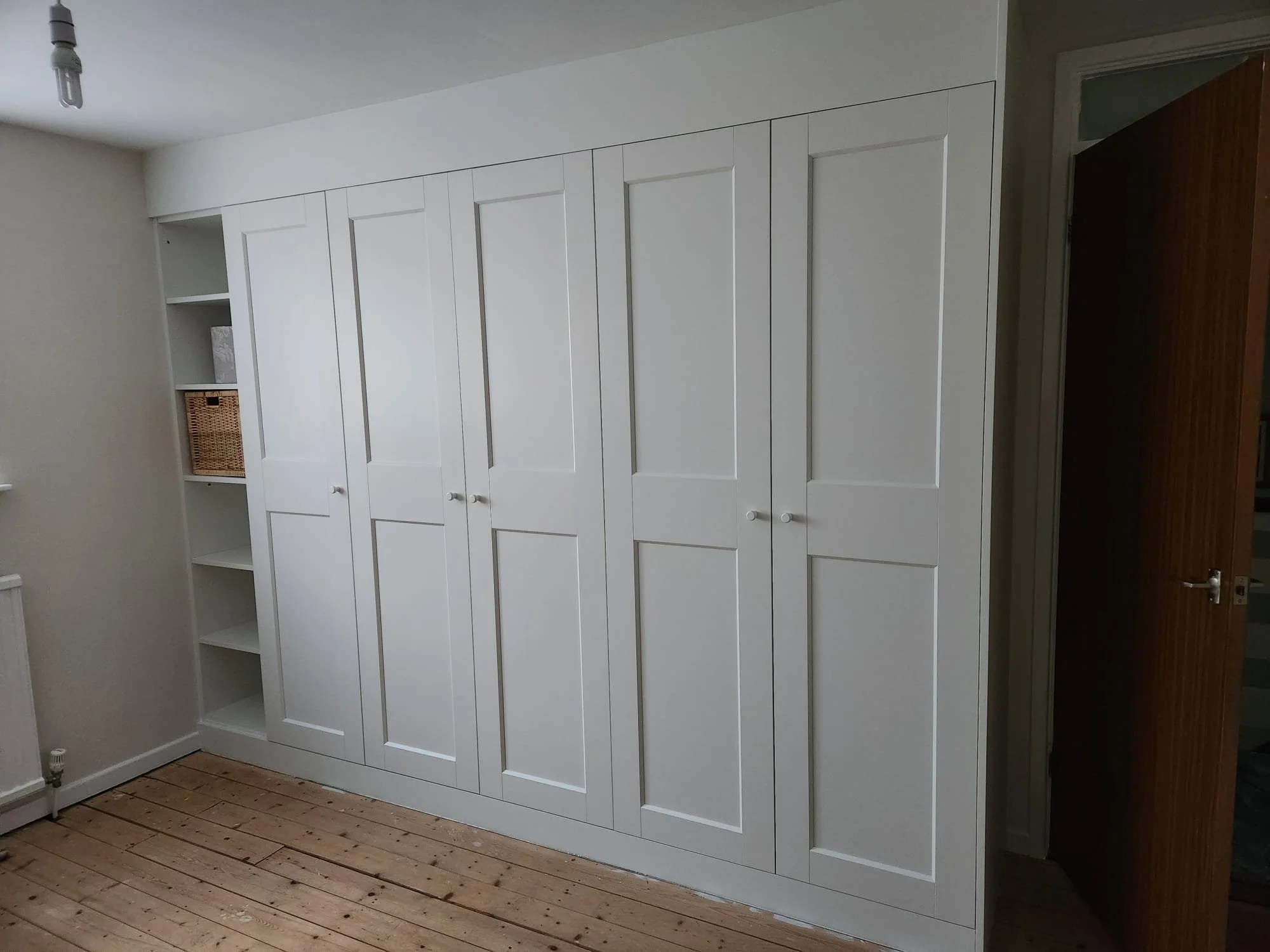 White built-in closet with five tall doors and an open shelf section on the left, in a room with a wooden floor and a light-colored wall.