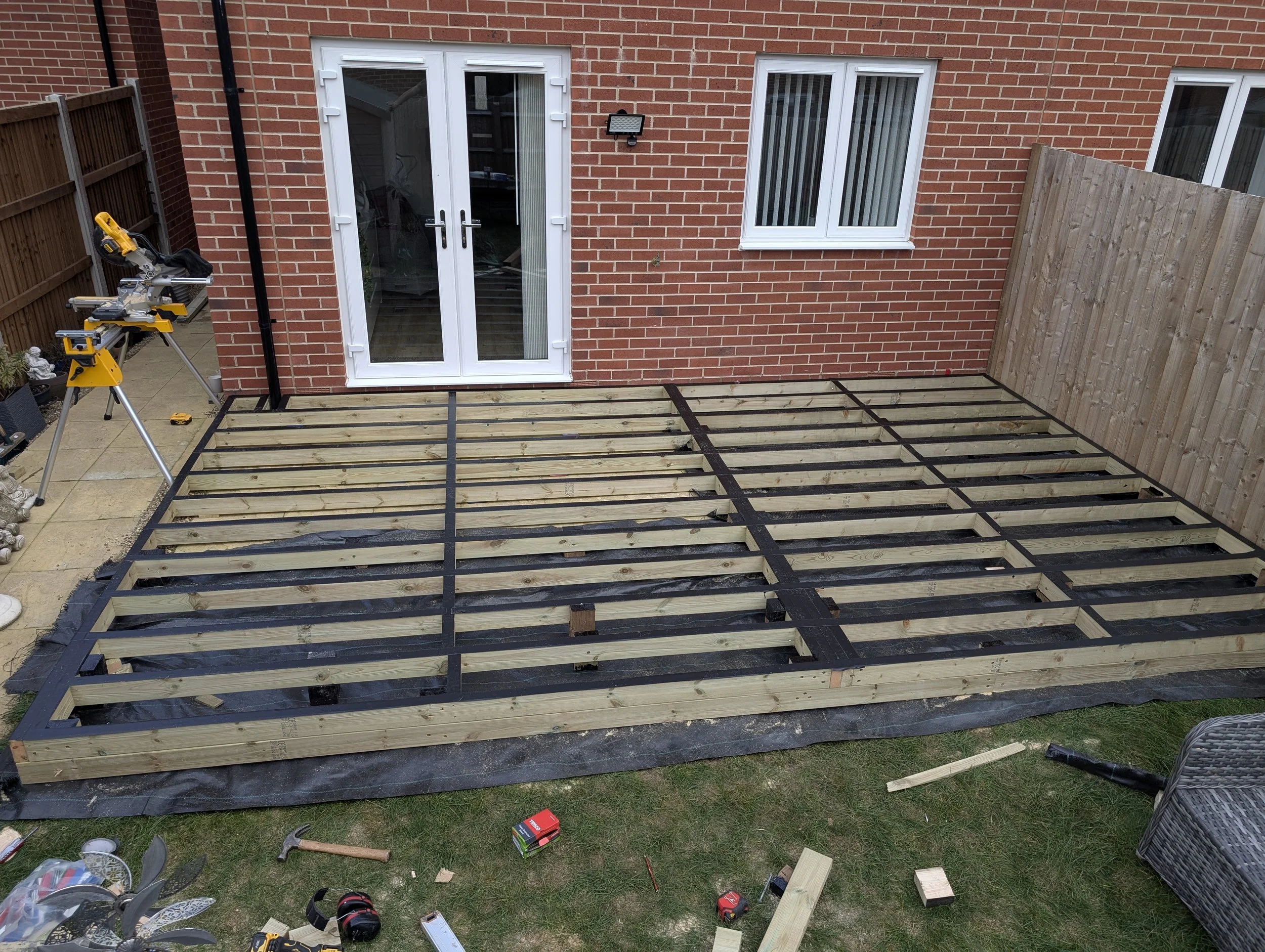 Construction of a backyard deck with wooden framing and support beams, next to a brick house with white double doors and windows, tools and supplies scattered around.