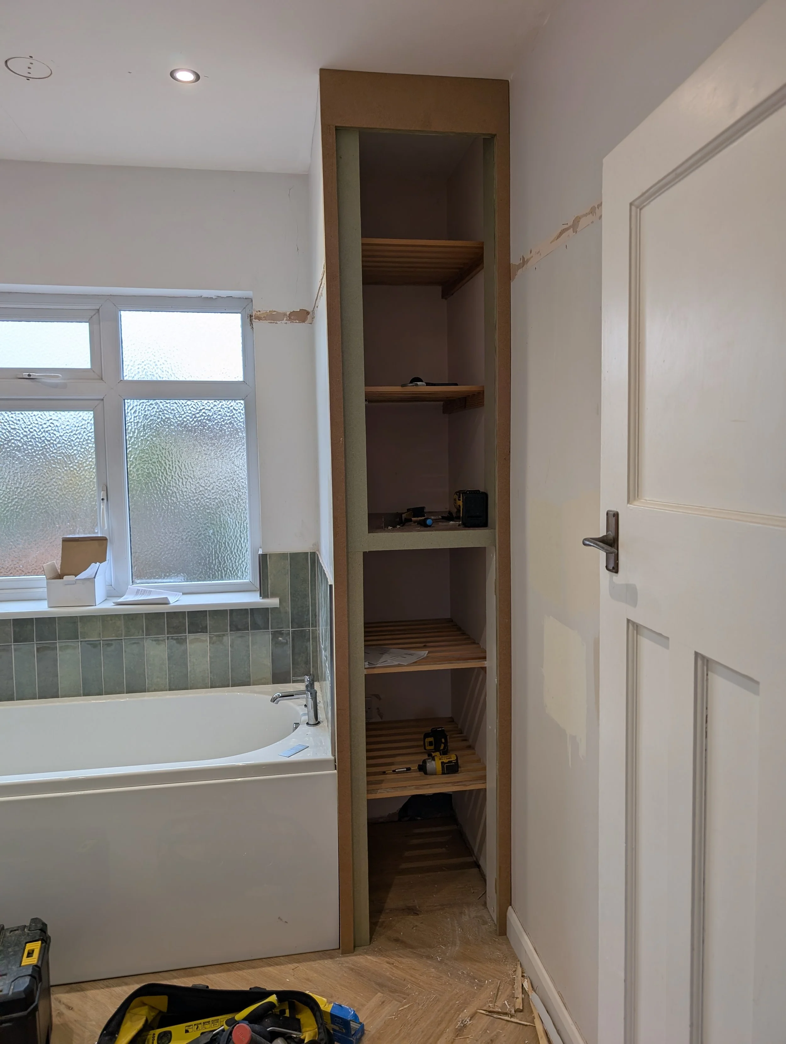 Inside a bathroom under renovation, featuring a bathtub near a frosted window, an unfinished shelving unit with tools, and construction materials on the floor.