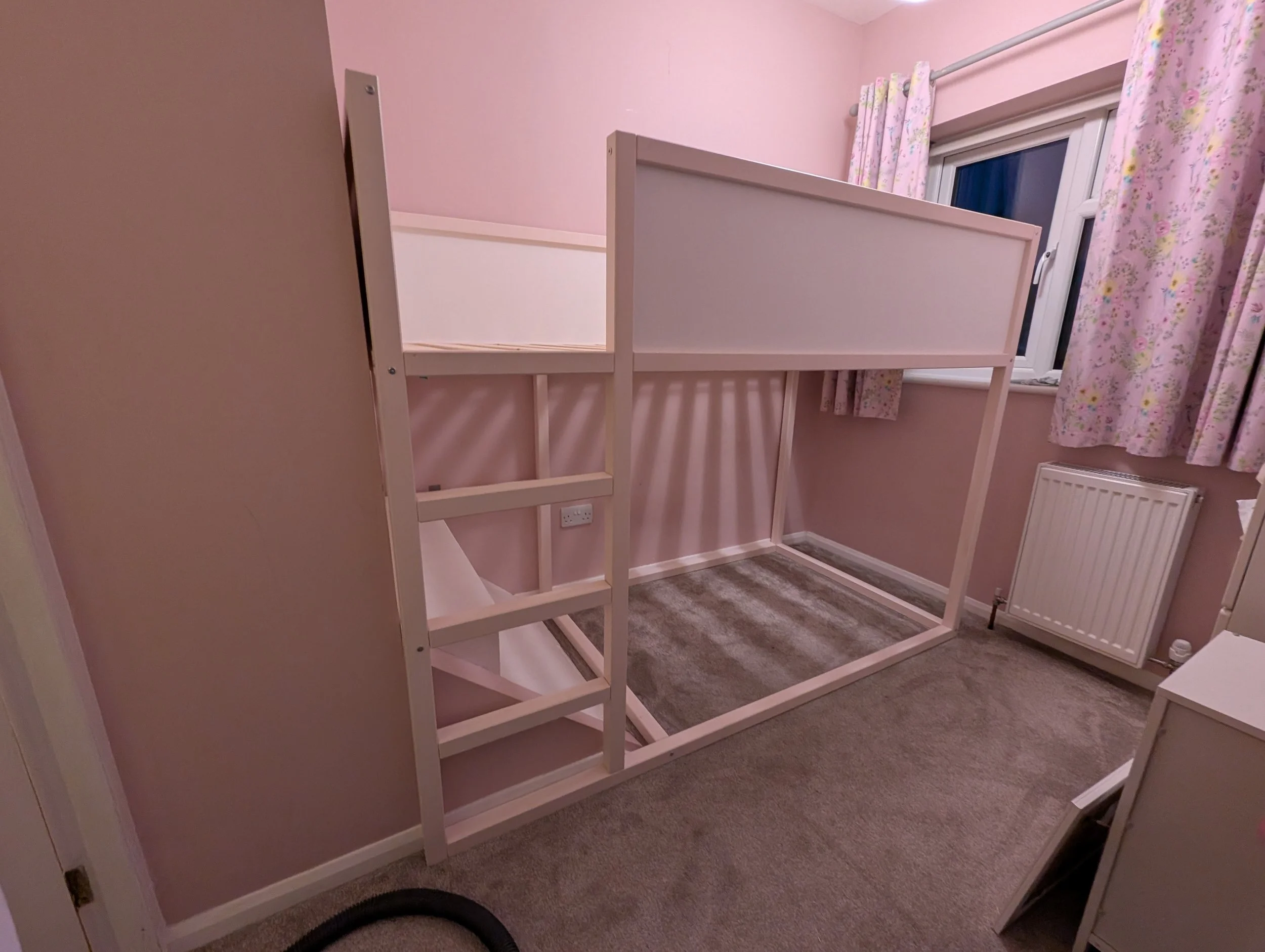 A room with pink walls, a window with pink floral curtains, a white radiator, and an unfinished loft bed frame with a small staircase.