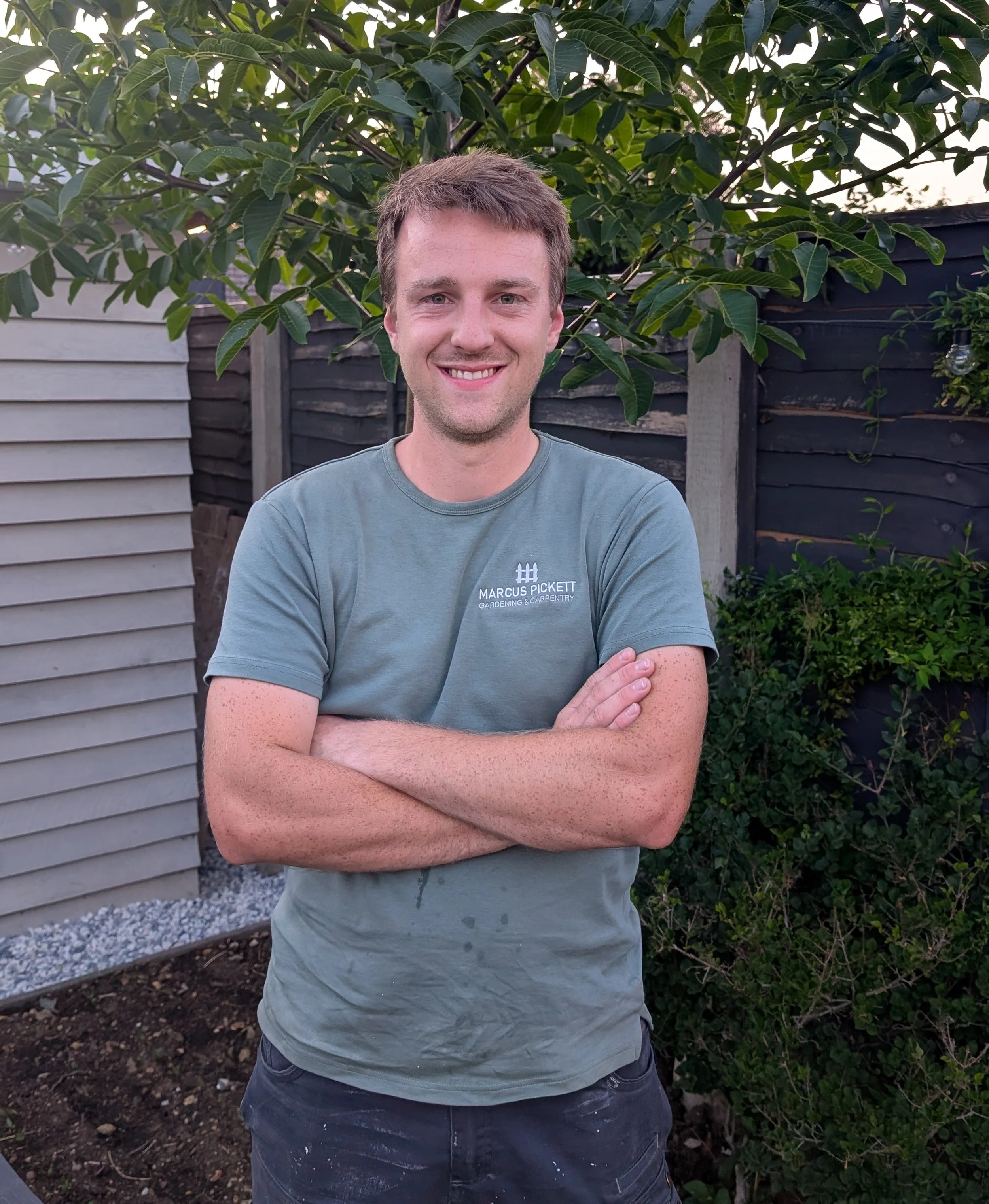 A young man with short brown hair standing outside in front of a tree with broad green leaves. He is smiling and crossing his arms. He is wearing a light gray t-shirt with the text 'Marcus Pickett Gardening & Carpentry' on it. The background includes a wooden fence, a garden bed, and part of a house.