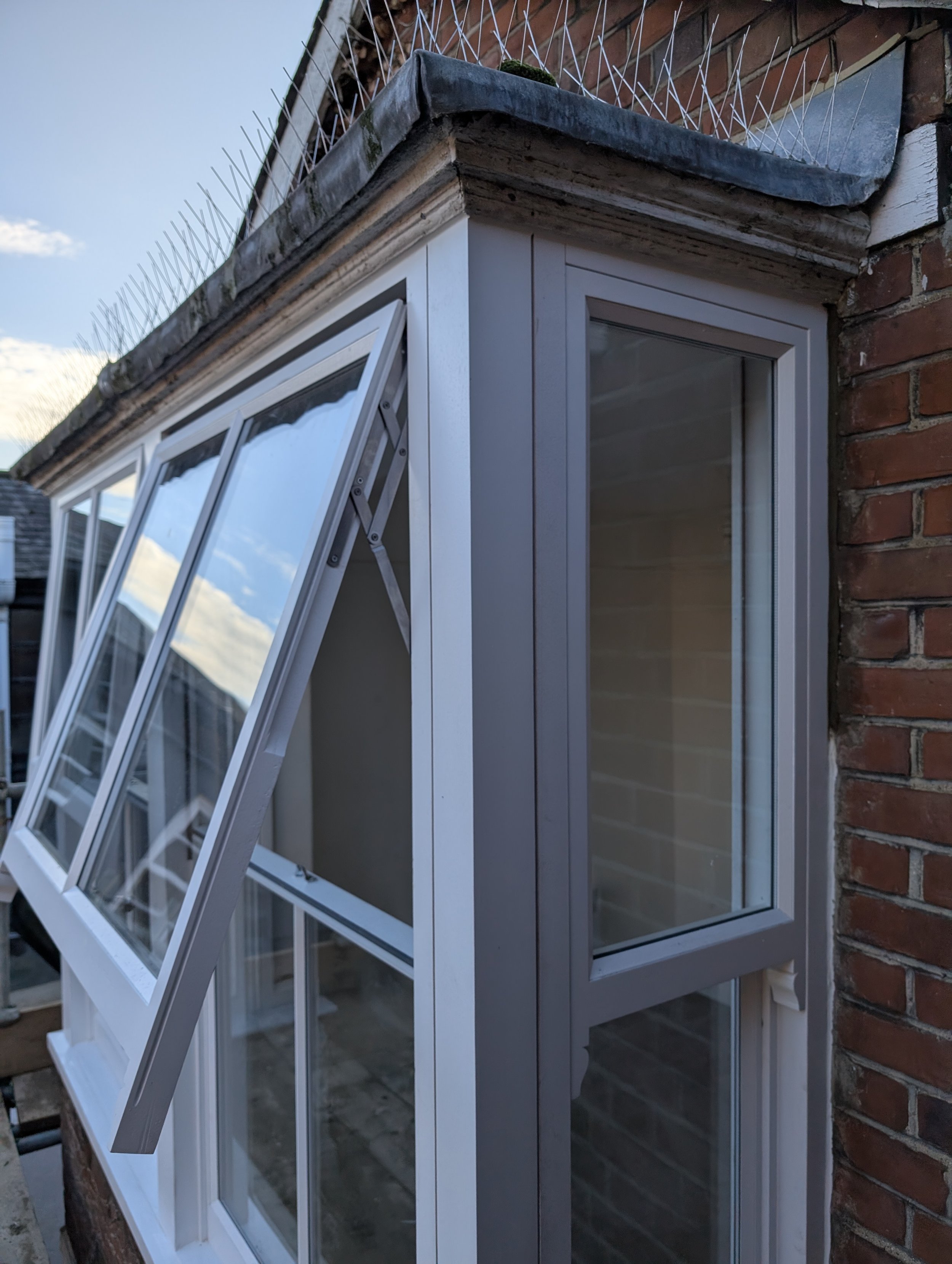 Close-up of a brick building corner with white window frames, some windows open, and a section of an outdoor metal fence at the top. The sky is blue with scattered clouds.