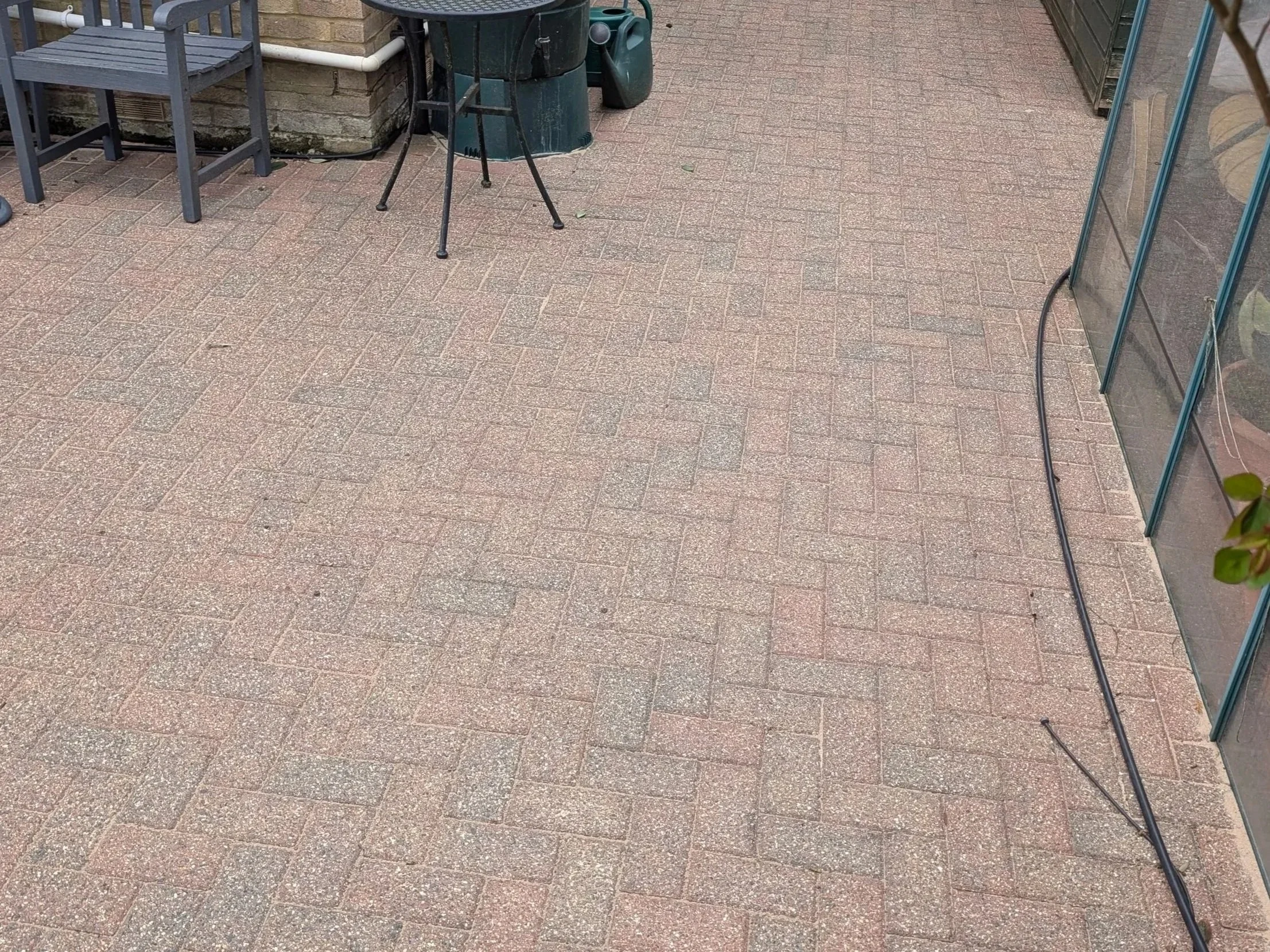 Outdoor patio with brick flooring, black chair, small green table, and a gardening tool near a brick wall and glass building.