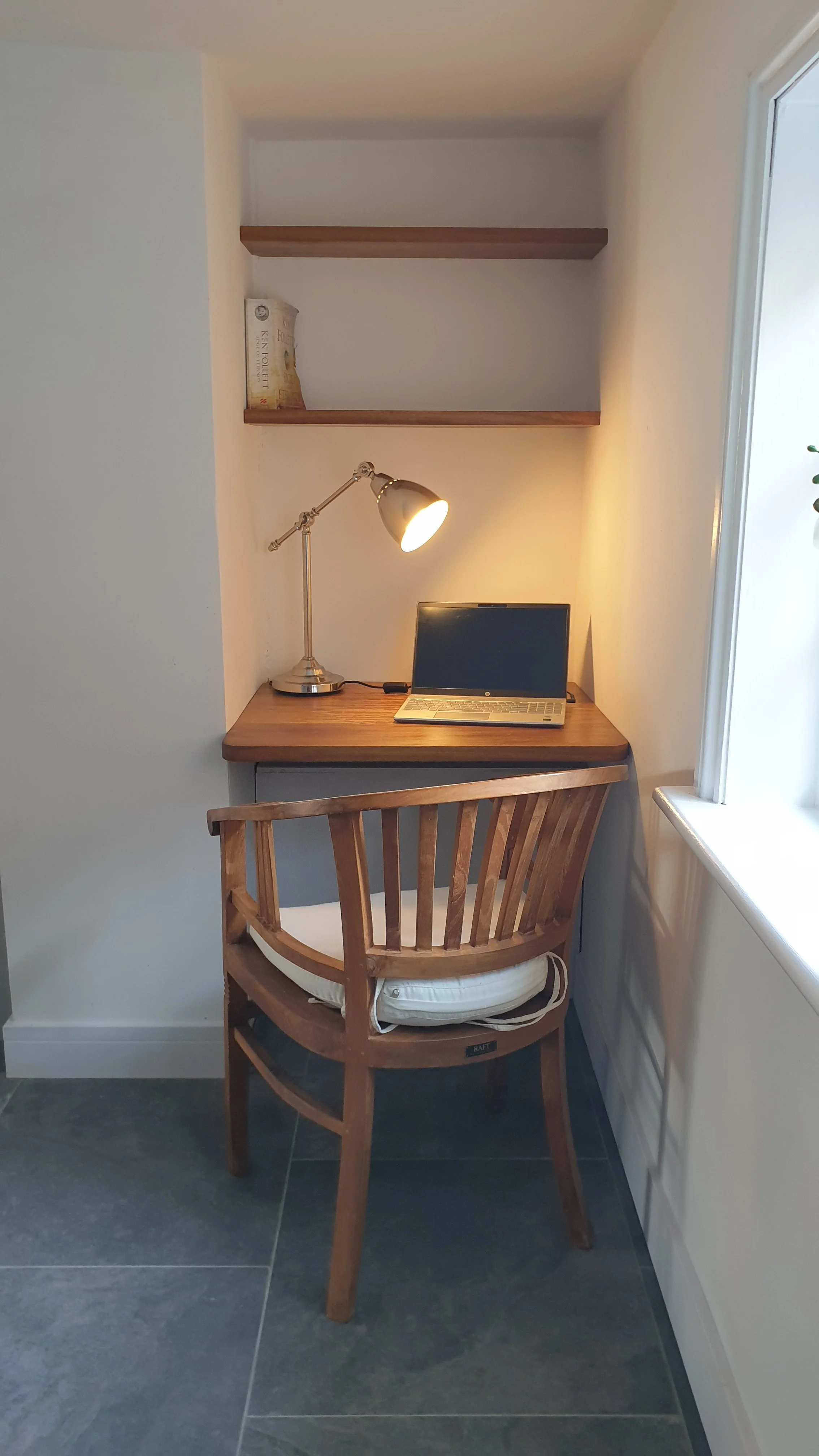 A small workspace nook with a wooden desk, a slatted wooden chair with a cushion, a laptop, and a desk lamp. There are two wooden shelves above the desk