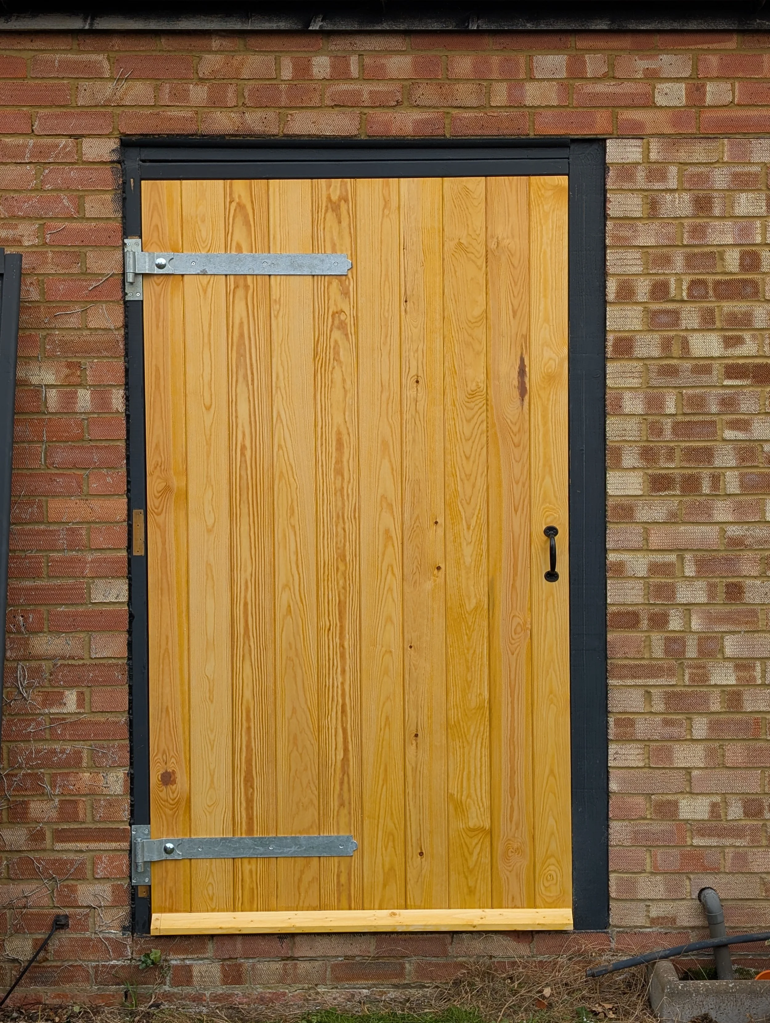 A wooden door with black trim installed in a brick wall, secured with metal hinges and a black door handle.