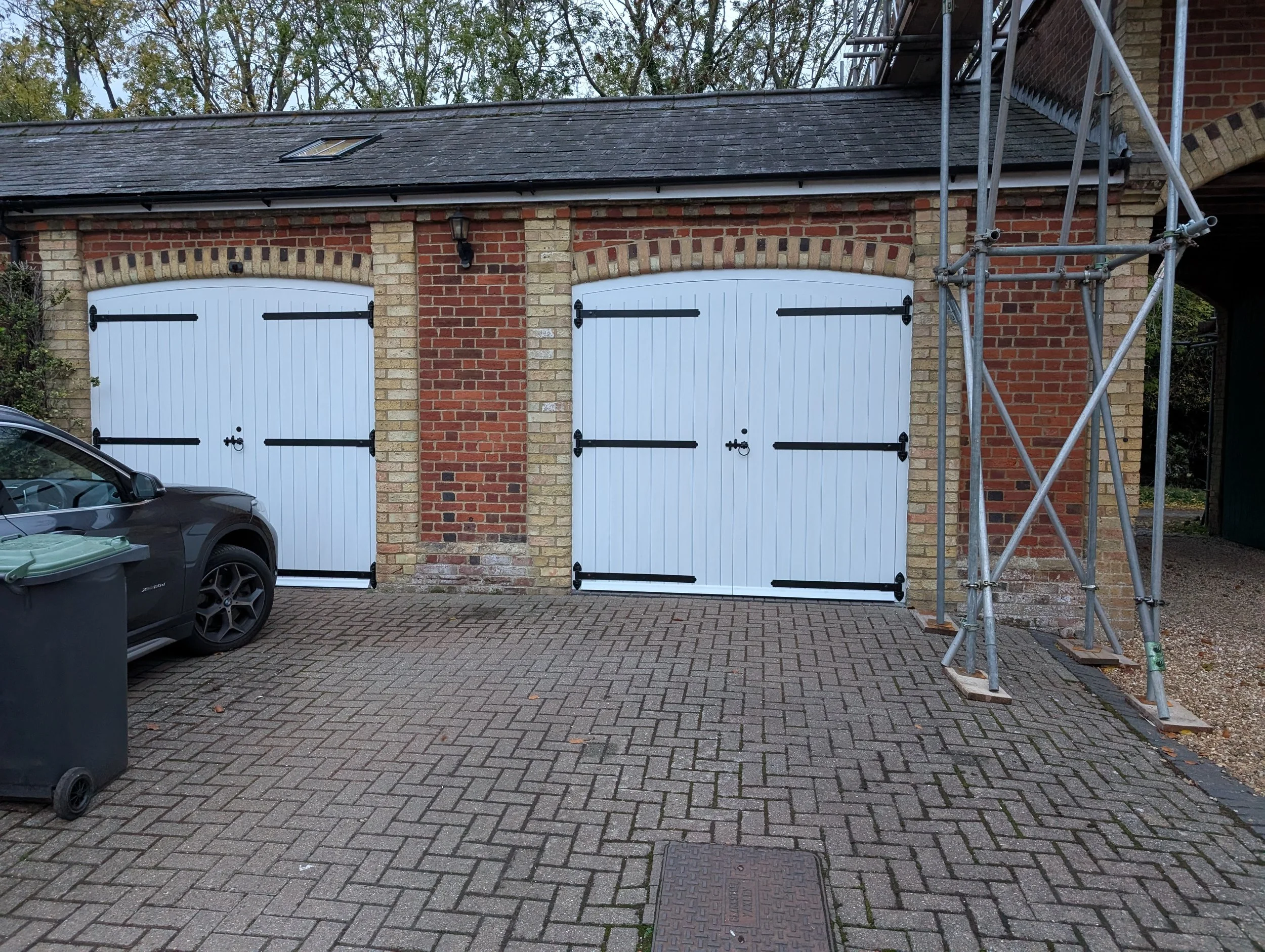 A brick garage with two closed white doors and black metal hinges, situated on a paved driveway. There is a gray car parked on the left, a black trash bin nearby, and scaffolding on the right side of the garage.