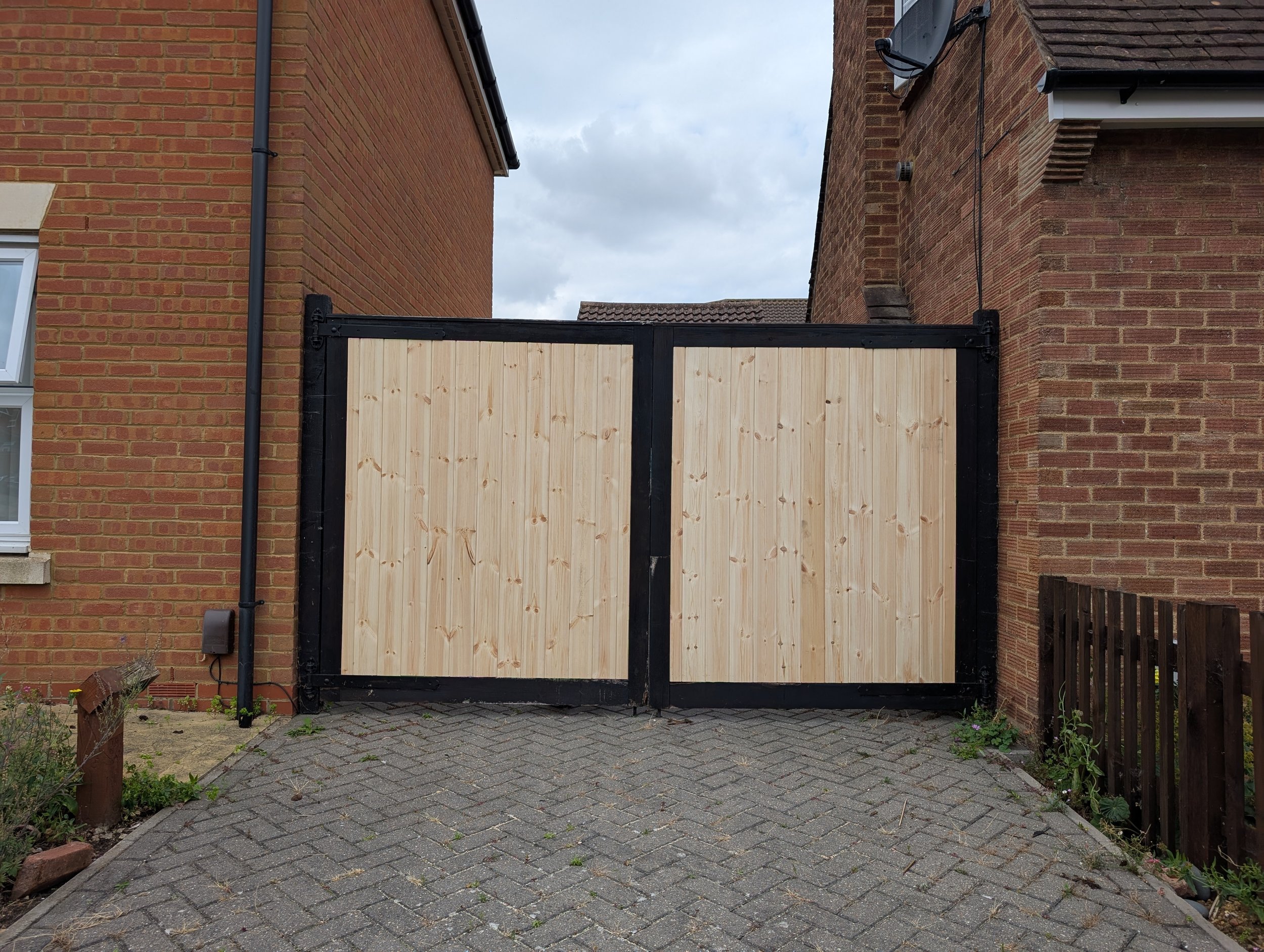A wooden gate with black frame between two brick houses, paved driveway, cloudy sky