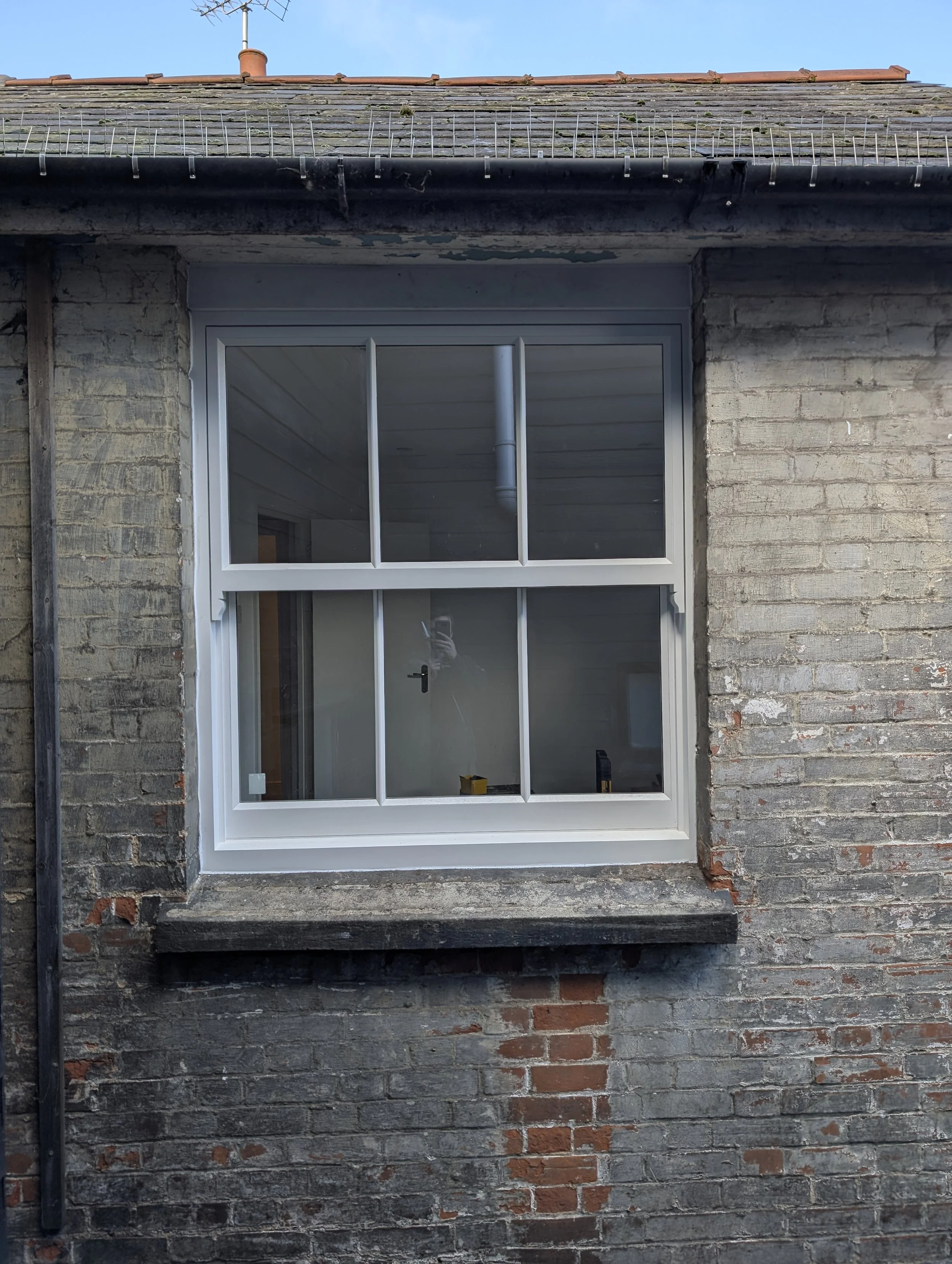 A newly installed white-framed, double-pane window on a brick exterior wall of a building. The interior shows a ceiling, temporary tools, and a person taking the photo.