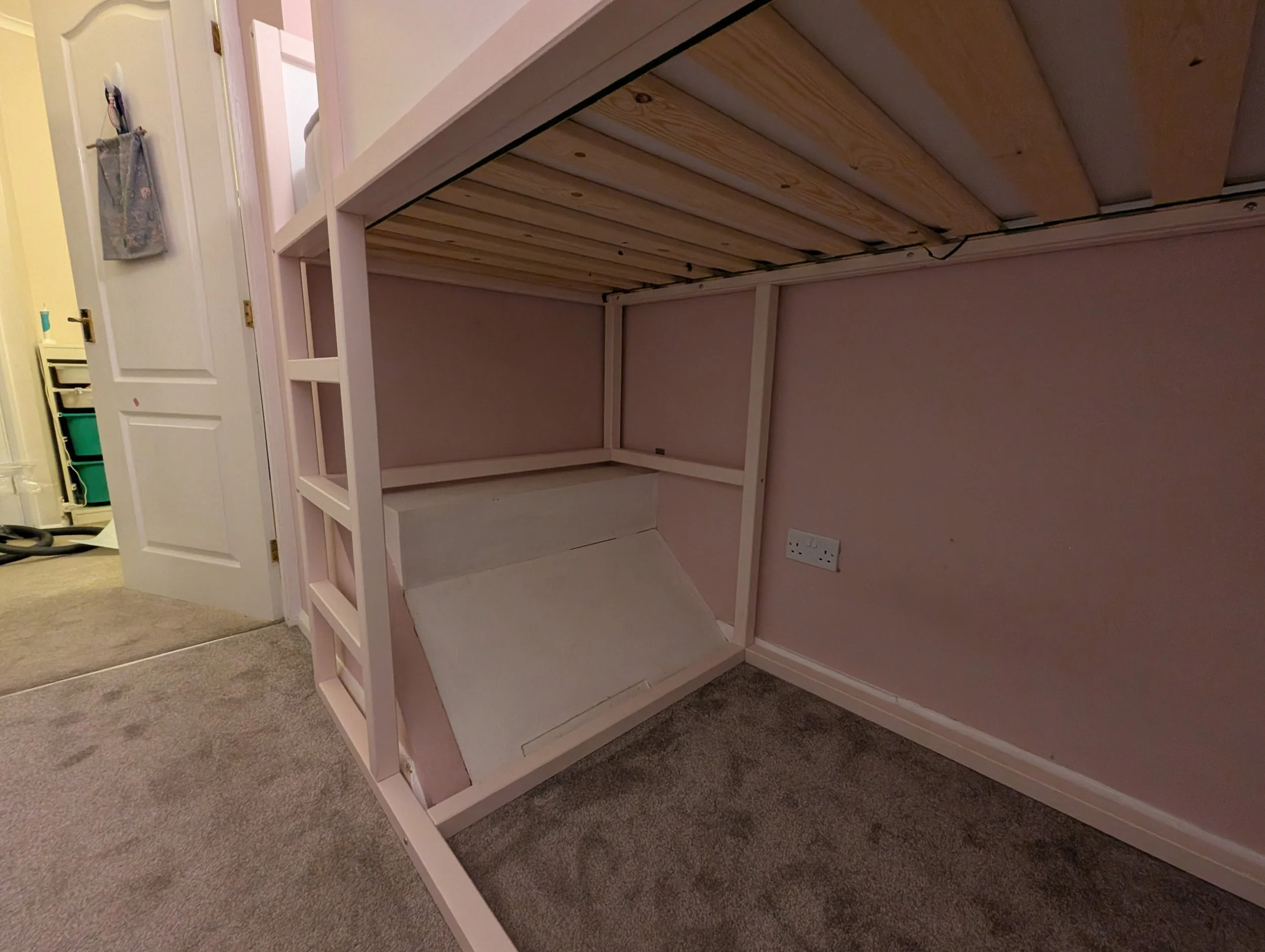 An unfinished pink-painted loft bed with a slanted front panel, located against a pink wall in a room with beige carpet. Part of an adjoining room is visible through an open door, with a green storage bin in the background.