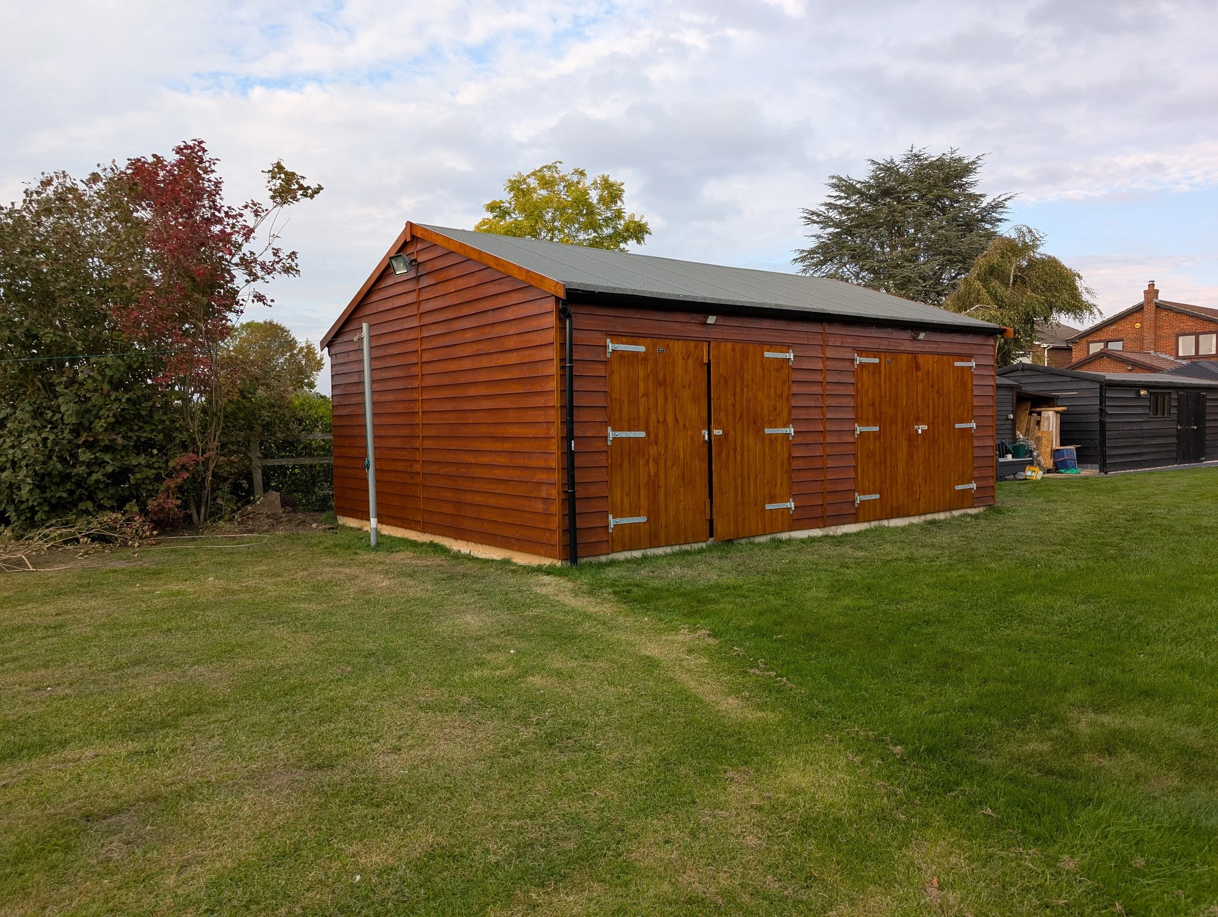 A wooden garden shed with a pitched roof, double doors, and a small window, situated on a lawn with trees and neighboring houses in the background.
