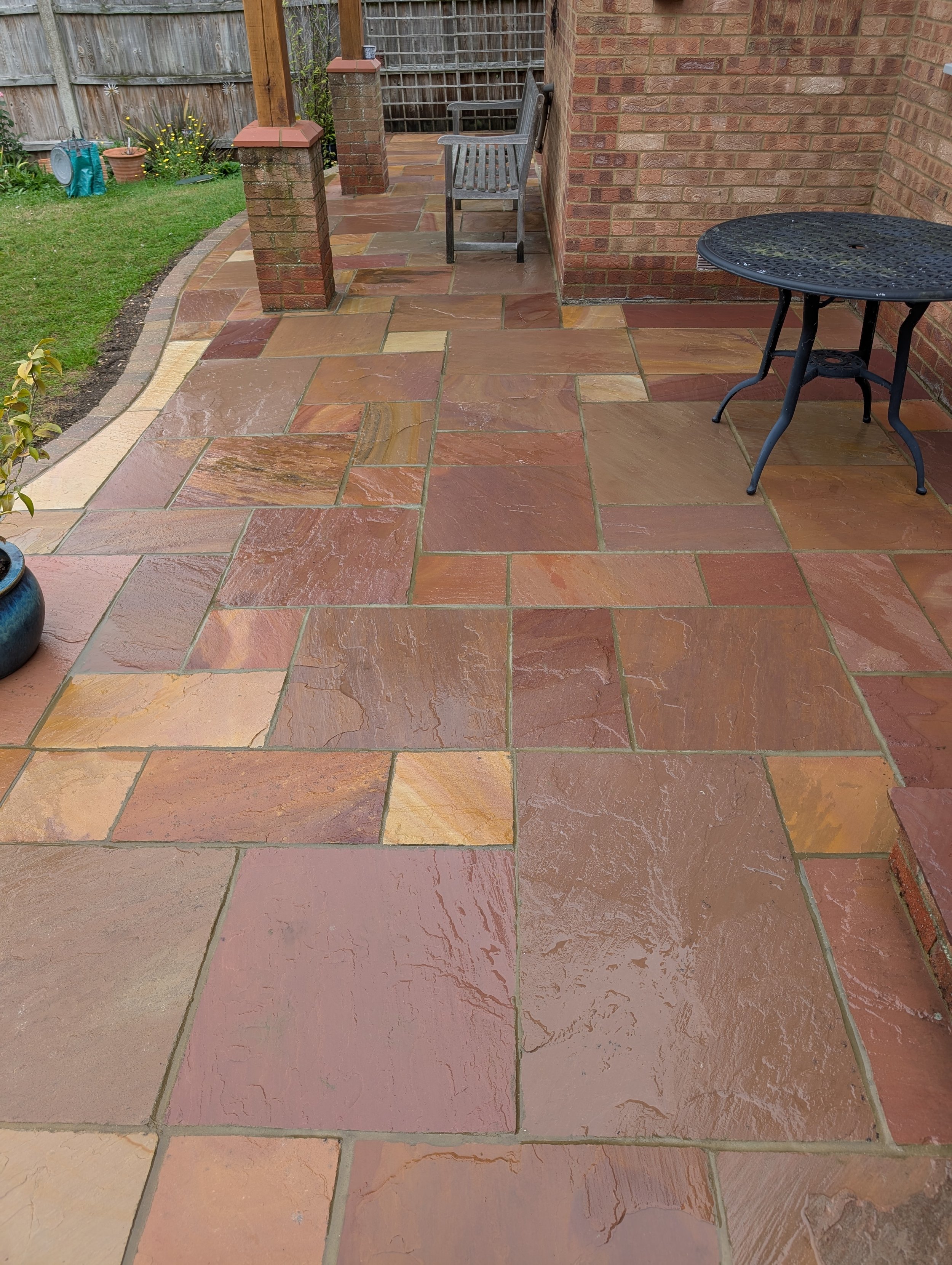 Wet multicolored stone patio with outdoor furniture, flower pots, and a lawn in the background.