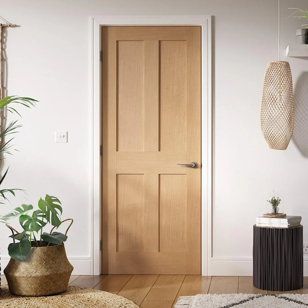 Interior view of a room with a closed light wood door, white walls, a black round side table with a plant, and woven pendant light; plants and decorative items are visible.