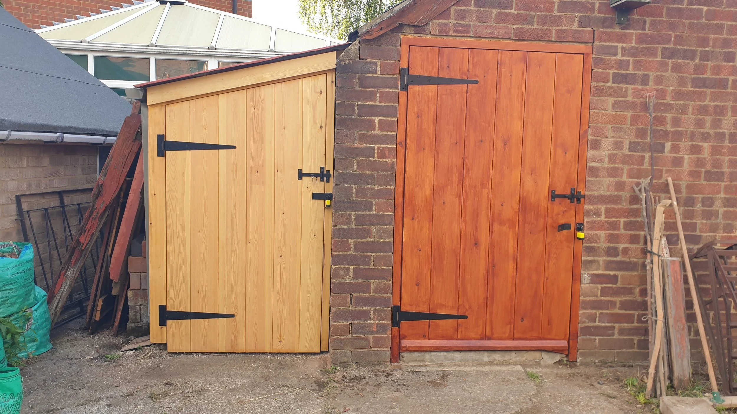 Two wooden garden sheds installed side by side on a brick wall. The shed on the left is lighter natural wood, with black hinges and a latch. The shed on the right is darker stained wood, also with black hinges and latch.