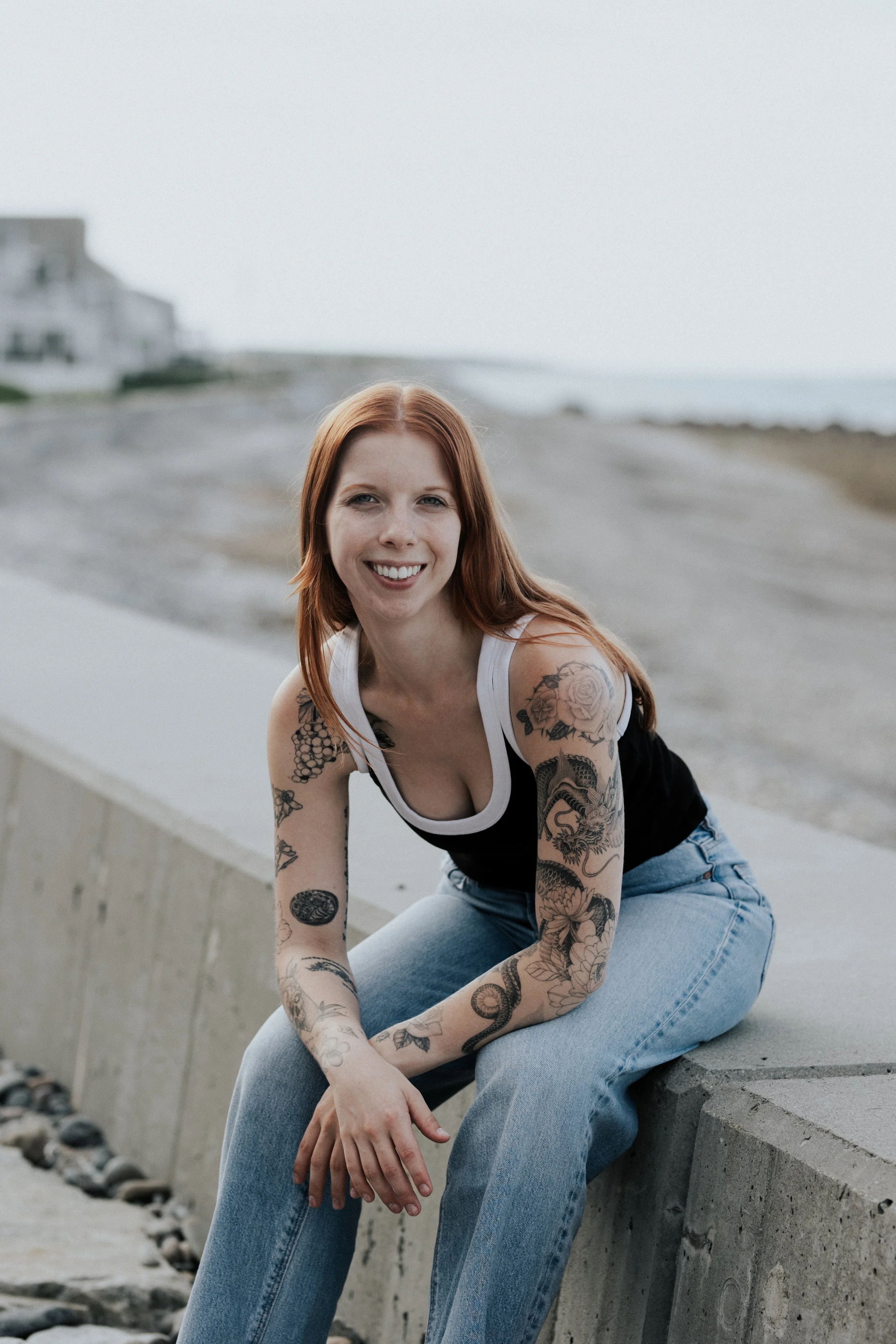 Person with long, red hair, tattoos, a black tank top, and lightwash jeans sitting on a cement wall in front of a blurred background