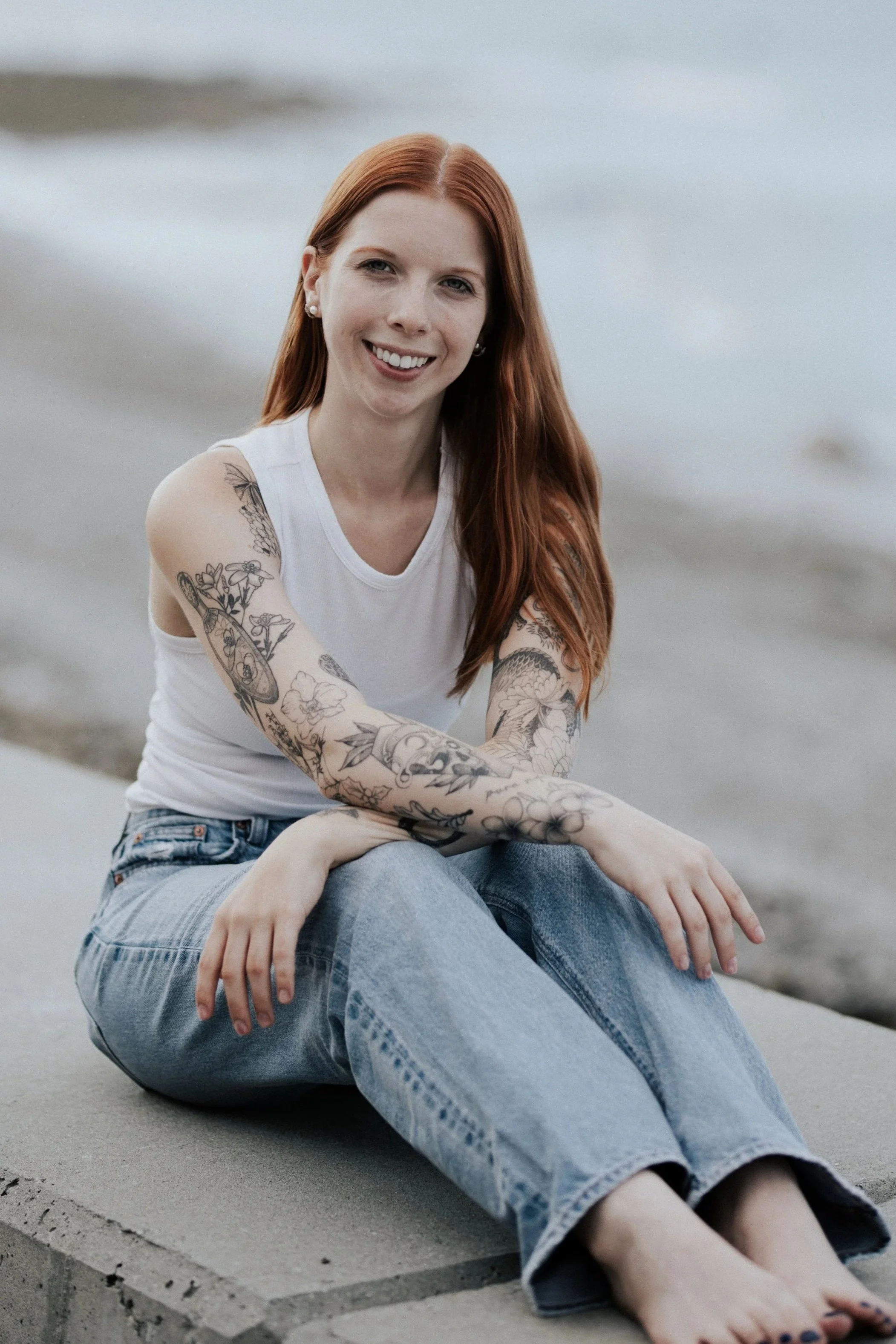 Kyra, a person with long, red hair, tattoos, a white tank top, and jeans, sitting with arms crossed over knees and smiling