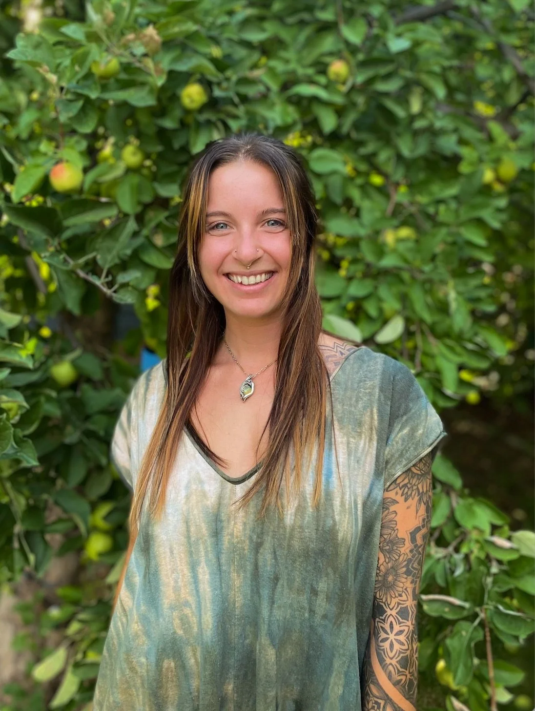 A woman with long brown hair and tattoos on her arm is smiling outdoors in front of a green leafy tree with small apples.
