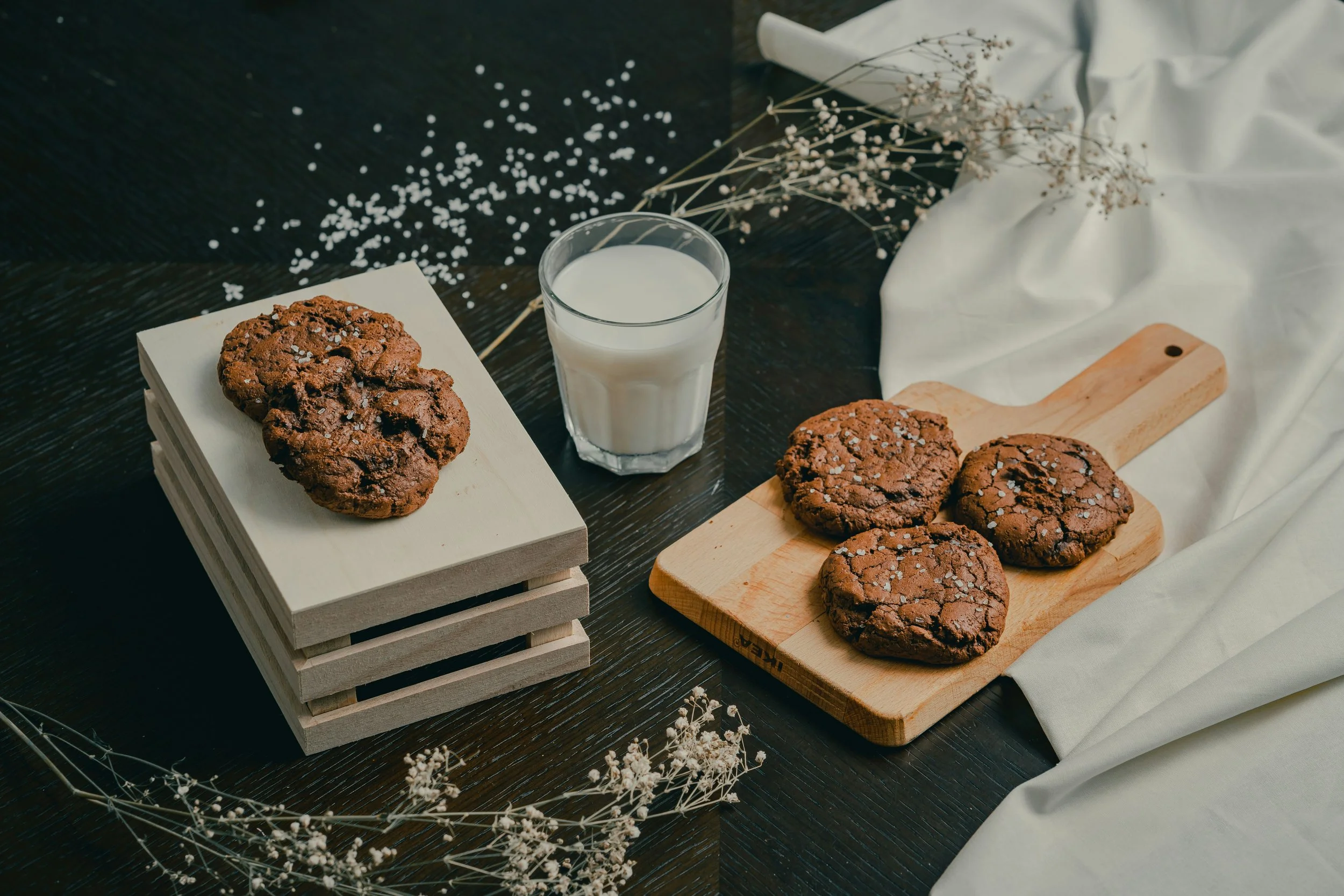 Chocolate cookies on a white crate, a glass of milk, and three more cookies on a wooden serving board, with white flowers and draped white fabric in the background.