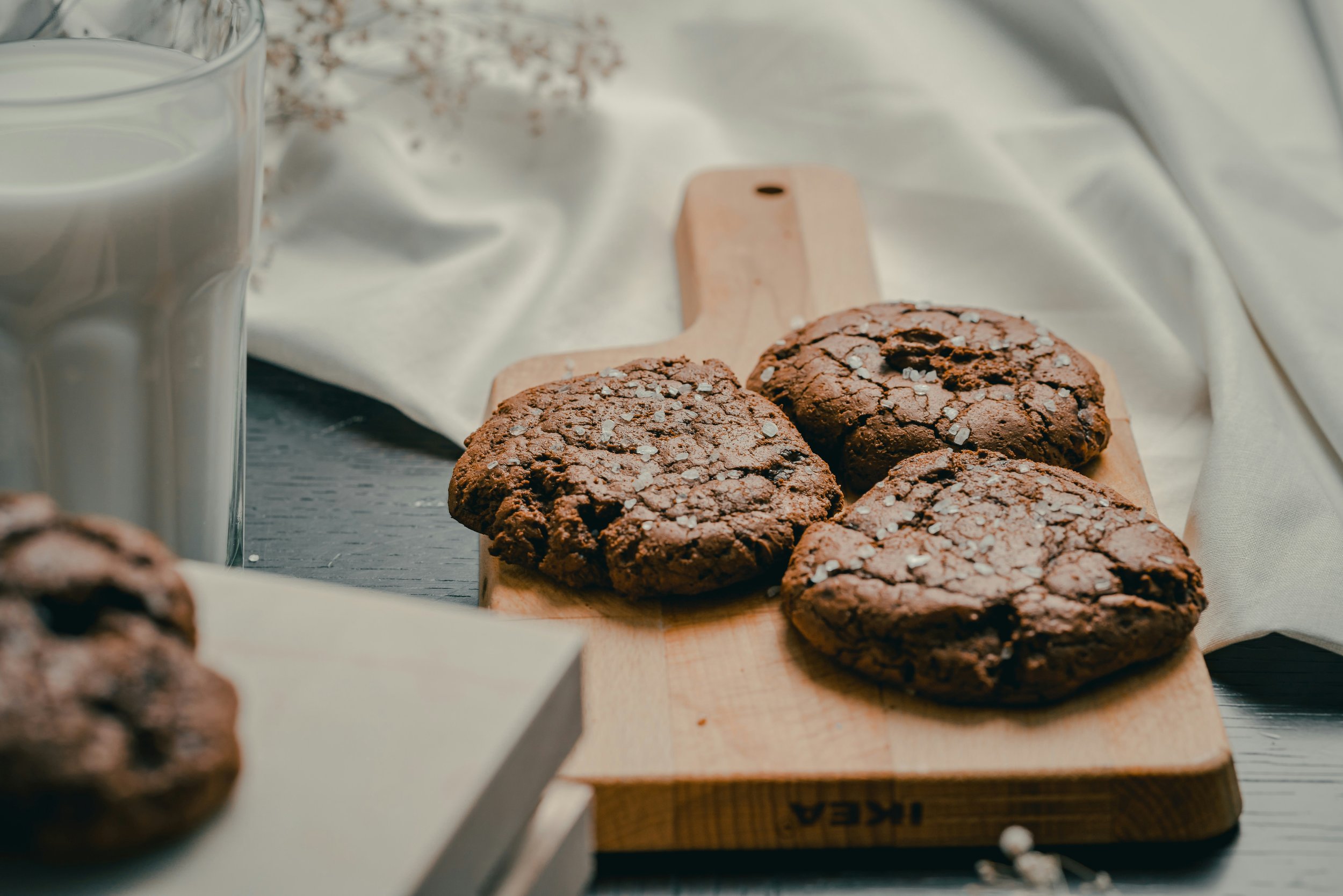 Chocolate cookies with sea salt on a wooden cutting board, glass of milk, and a white cloth.