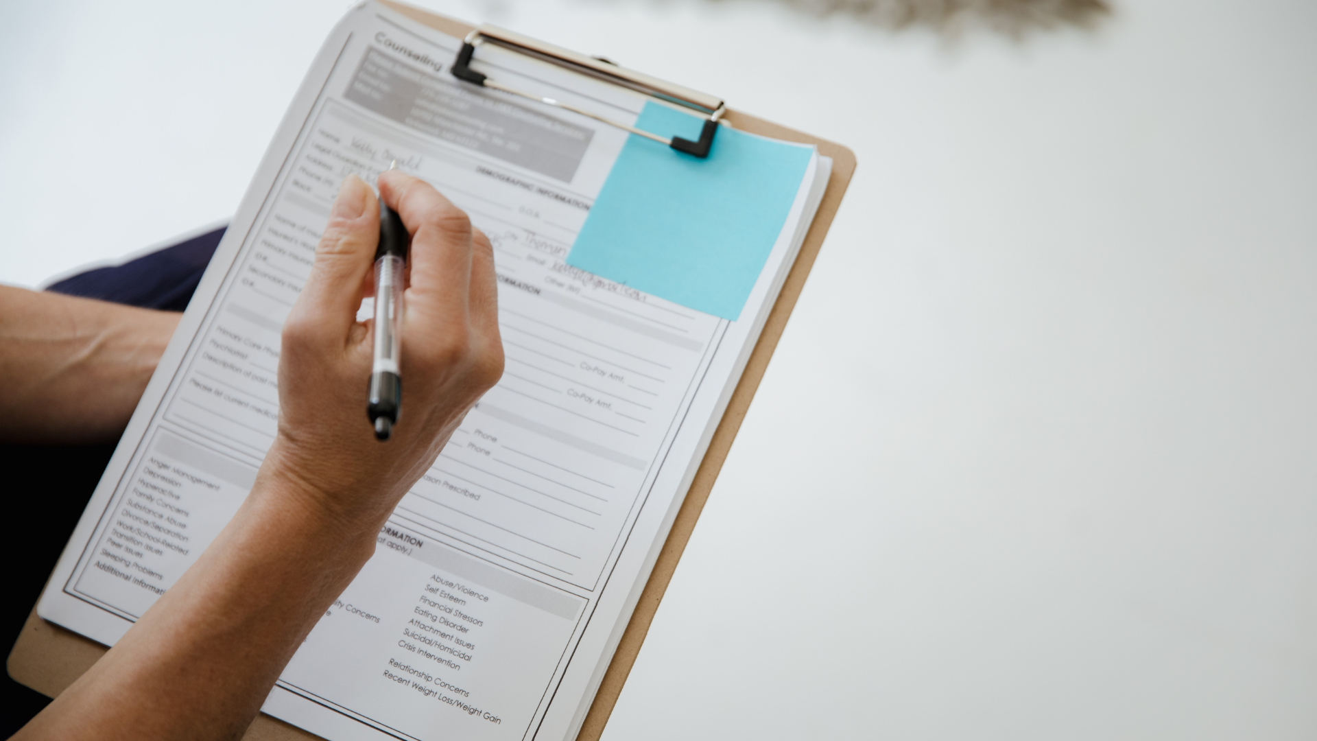 Eine Person schreibt auf einem Diagnostikbogen, der auf einem Pinnwandclip befestigt ist, auf einem Klemmbrett. Es befindet sich ein blauer Notizzettel auf dem Bogen.