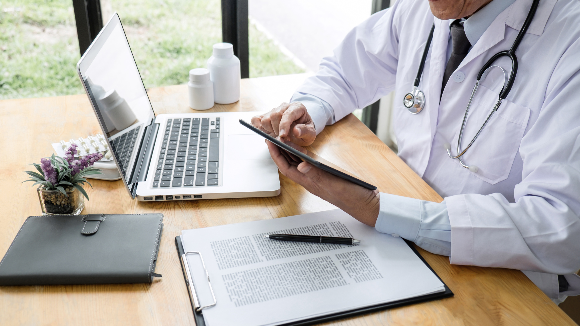 Arzt sitzt an einem Tisch mit einem Laptop, einer Pflanze, Medikamentenflaschen, einem Klemmbrett mit Dokumenten, einem Stift und einem Tablet.