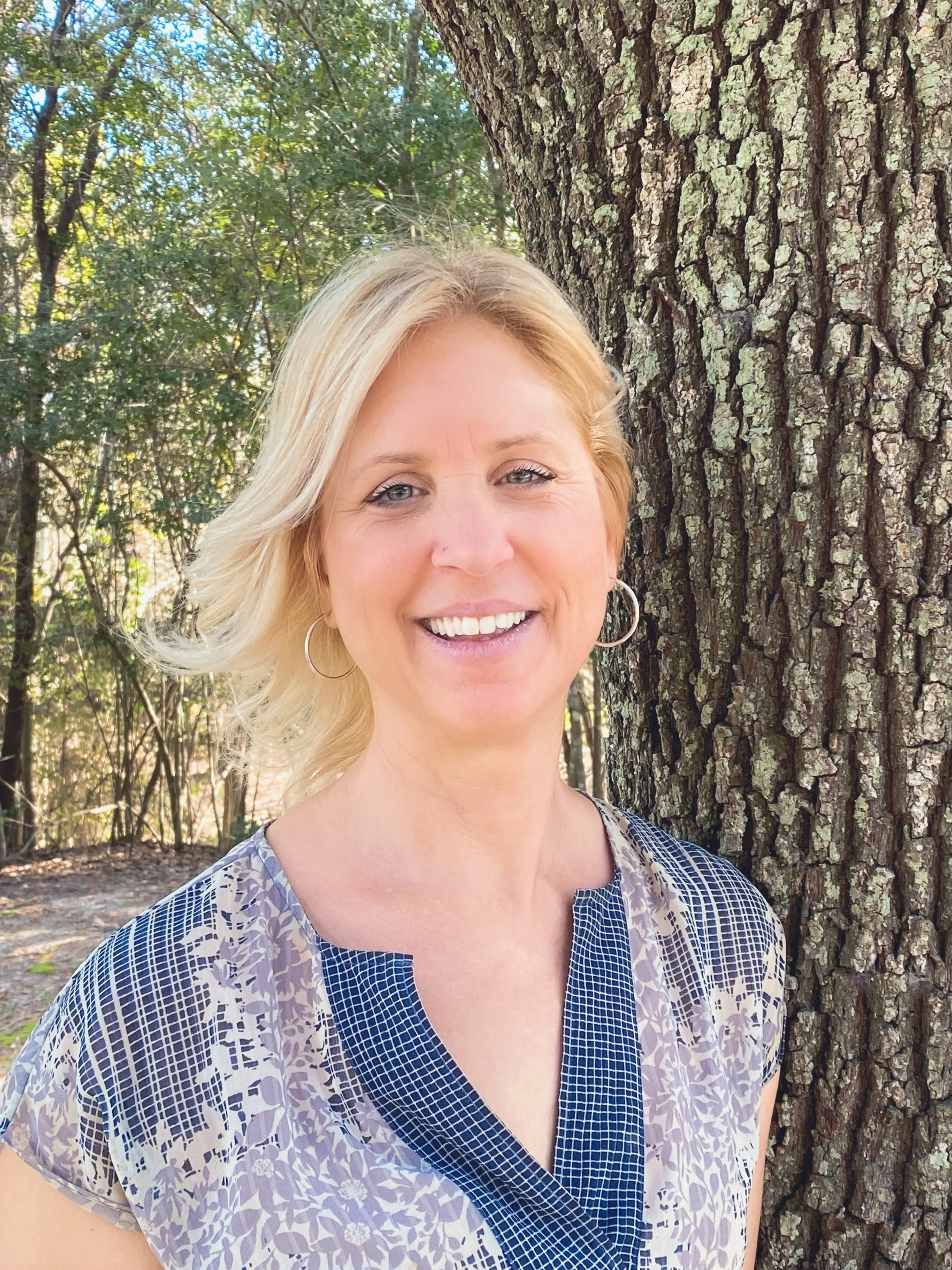 A smiling woman with blonde hair and blue eyes standing outdoors near a large tree trunk, with a background of green foliage and sunlight filtering through the trees.