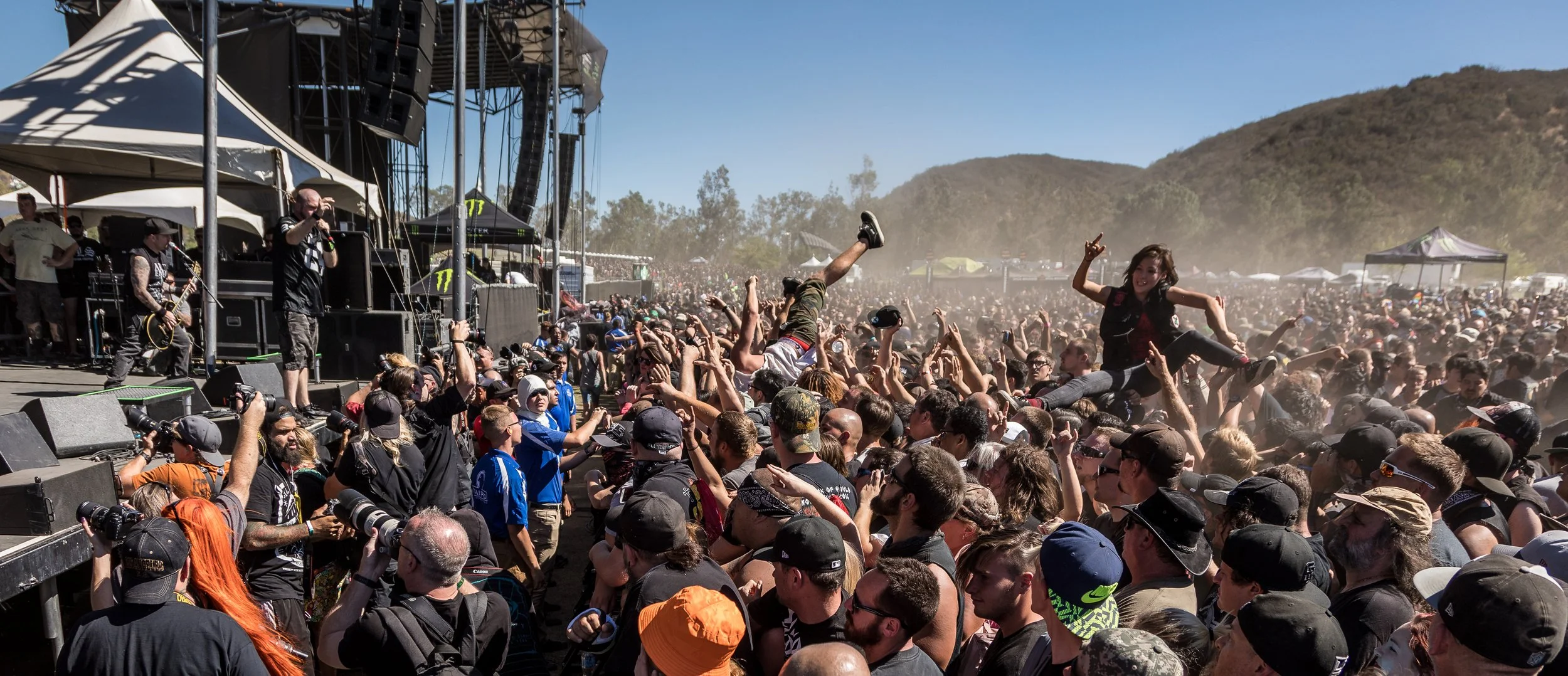 The band, on stage at the left, plays on while a couple people crowd surf on a bright sunny day