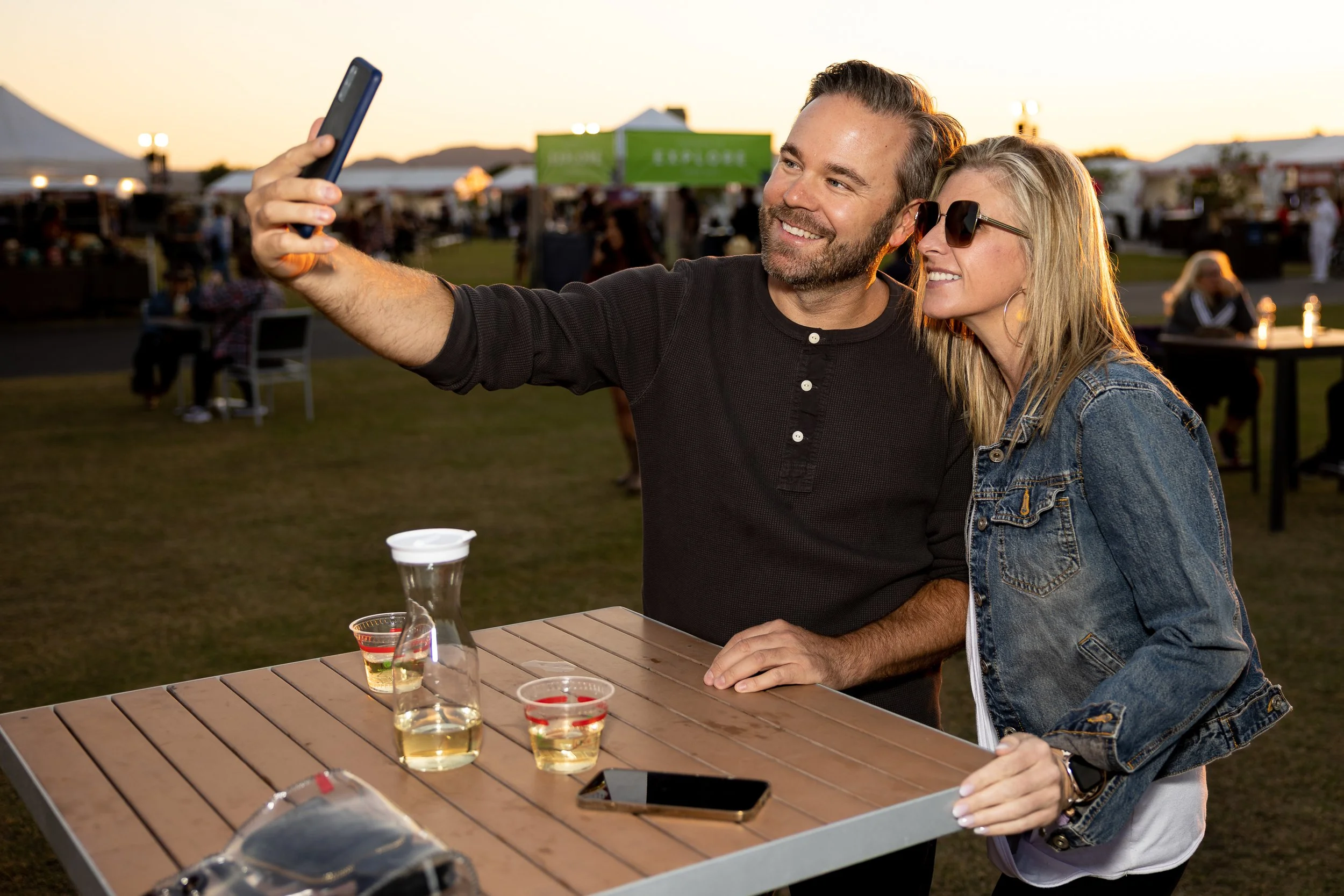 A man and woman are at a table during an outdoor event, taking a couples selfie