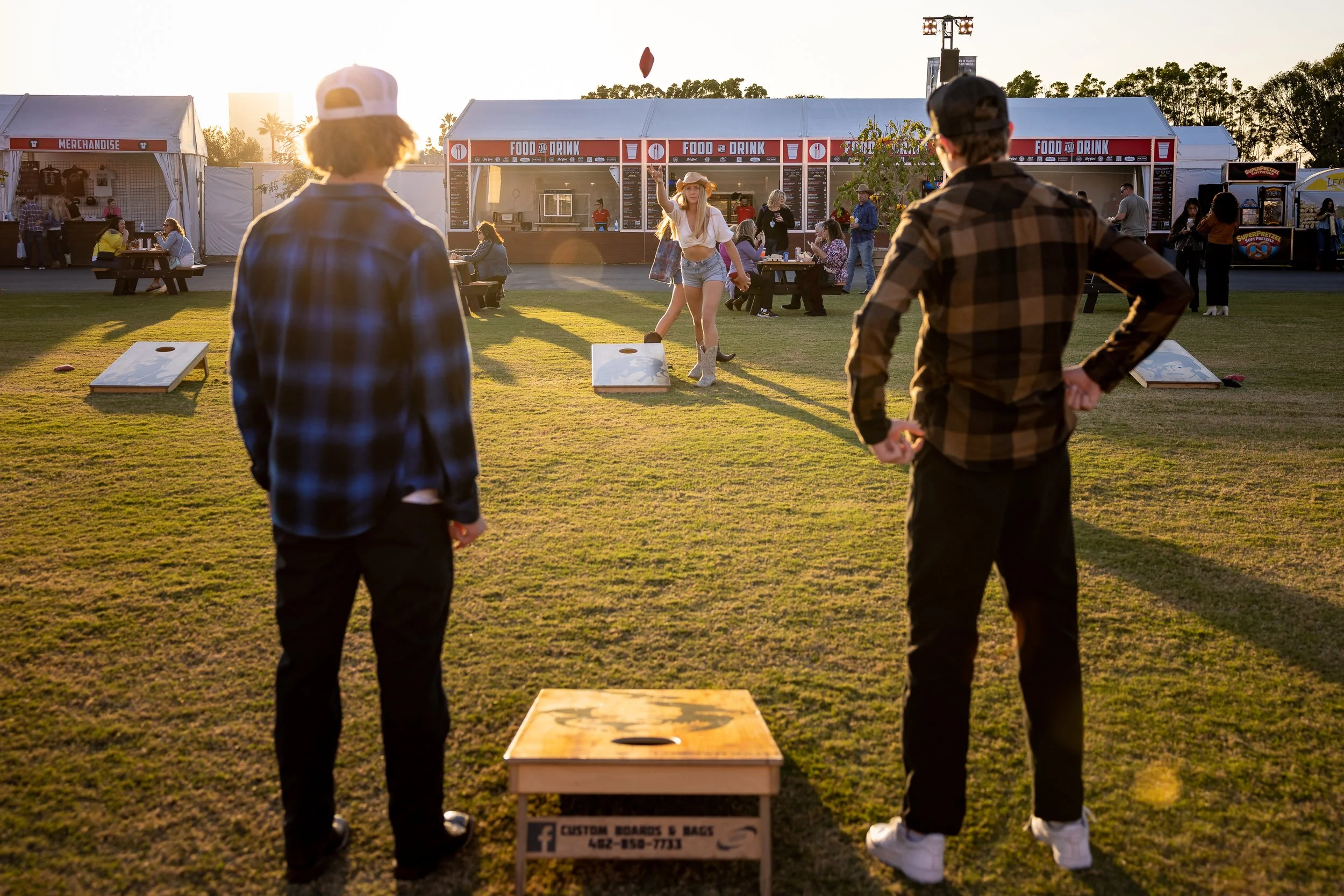 A woman throws a beanbag towards the camera, with a food and drink stand behind her as part of a game. Two men in plaid shirts stand in the foreground facing her