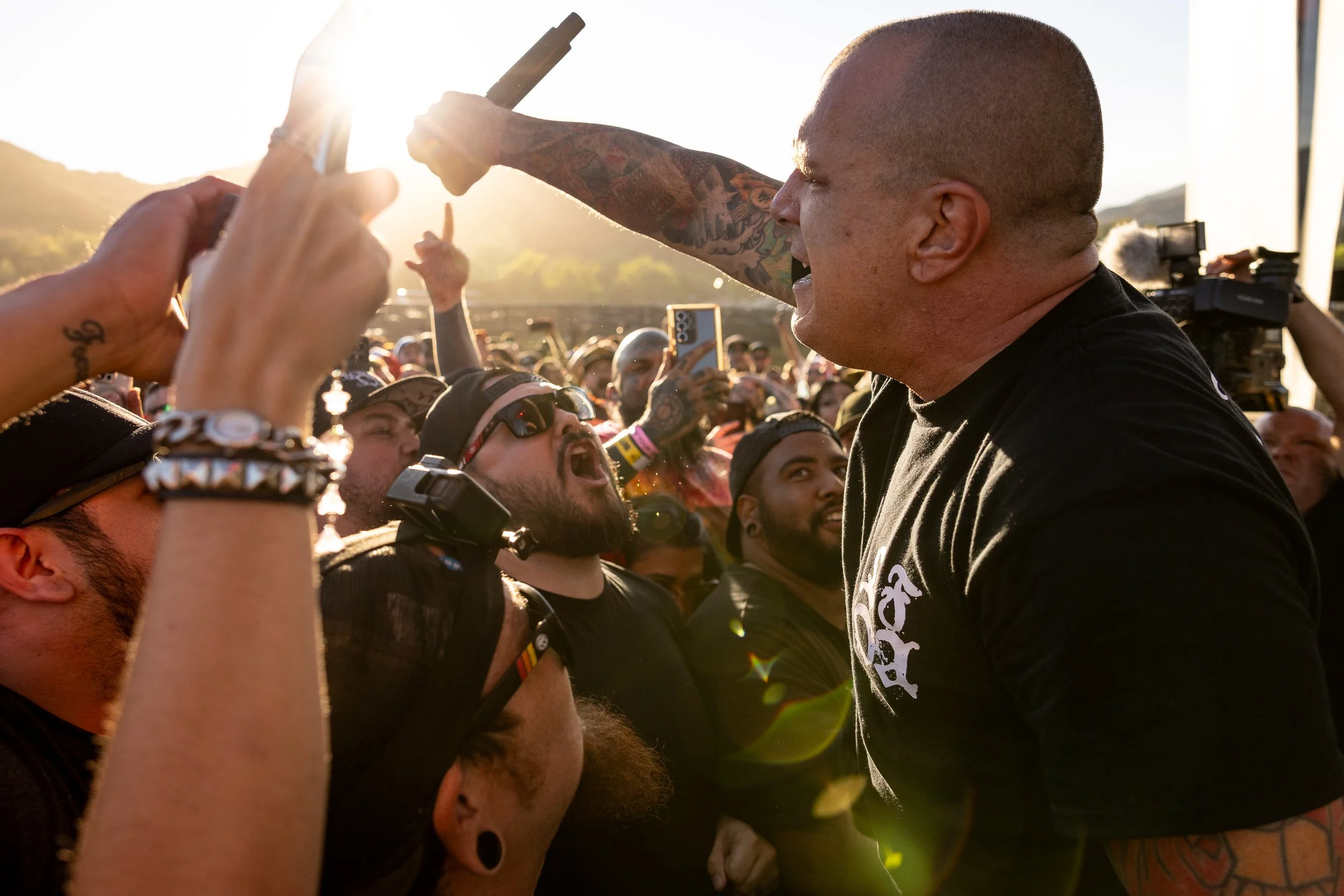 A tattooed, bald, male singer leans into the crowd, holding his microphone above them as they sing, with the sun bursting between his hand and he crowd's outstretched hands