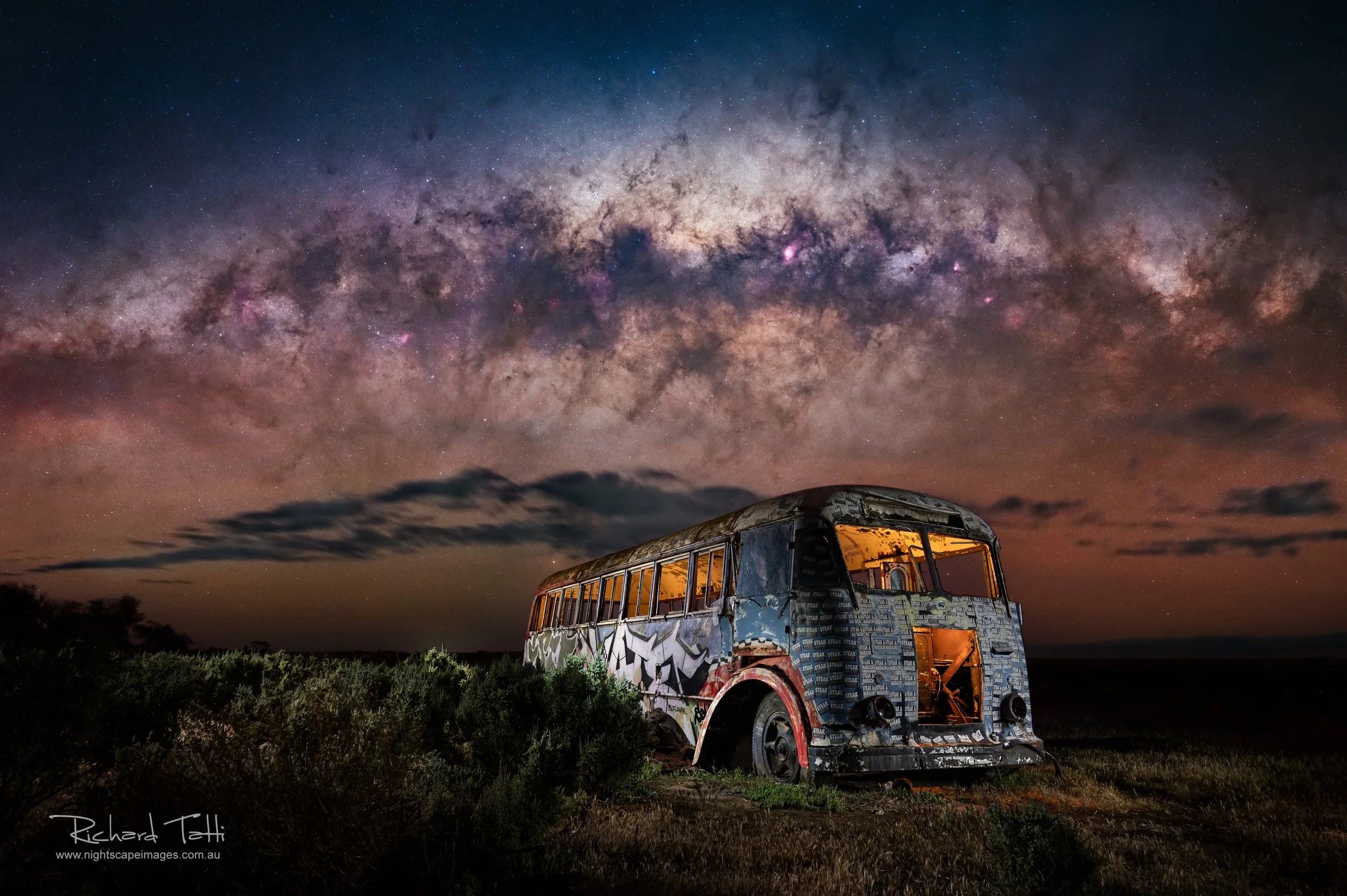 a broken down old bus, covered in graffitti, with warm light inside and the Milky Way in the sky above