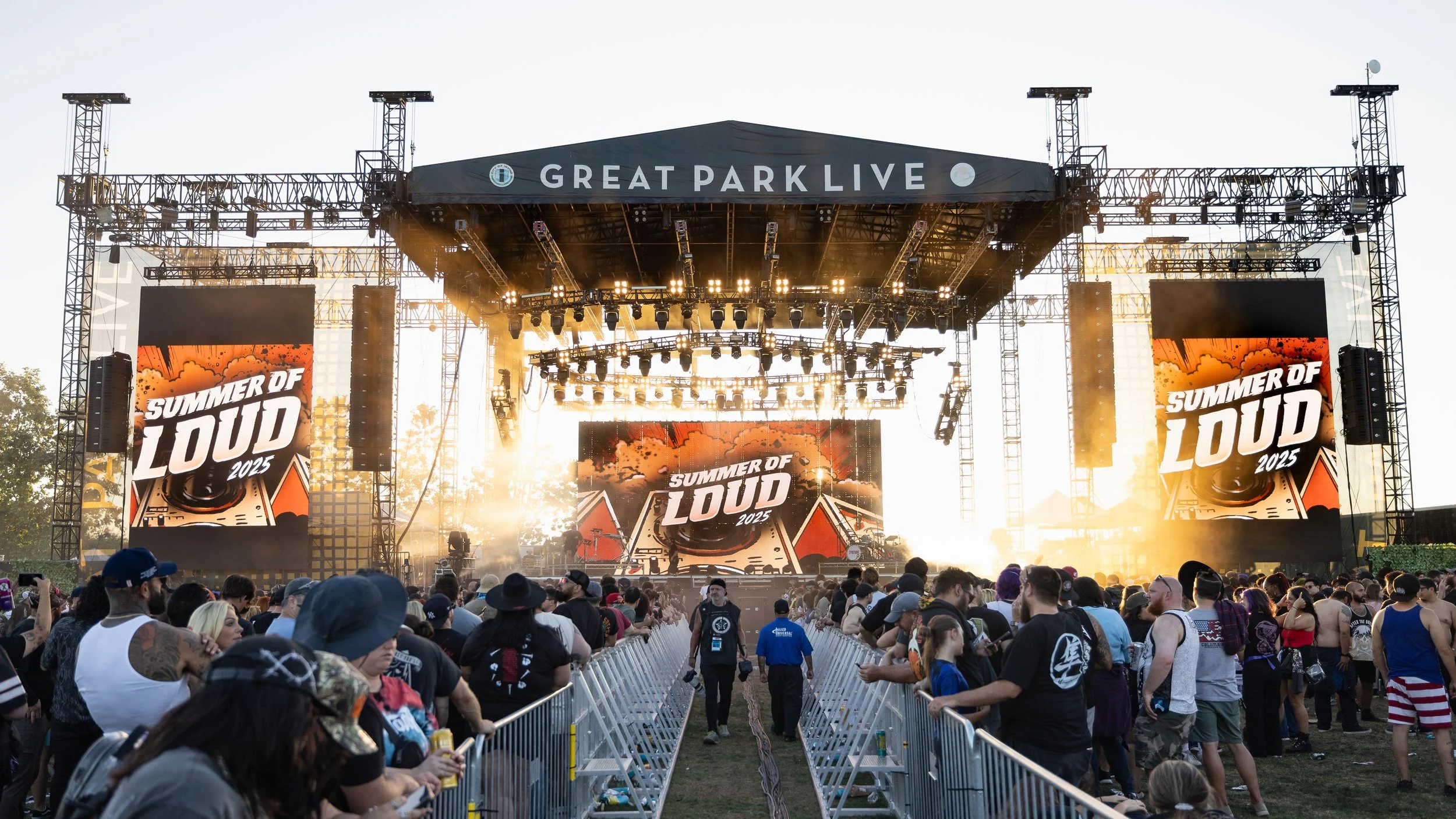 Like the parting of the seam tow fences run down the middle of the concert venue, leading to the stage. On either side of the fences, the crowd watches the band, as a photographer walks towards the camera right up the middle