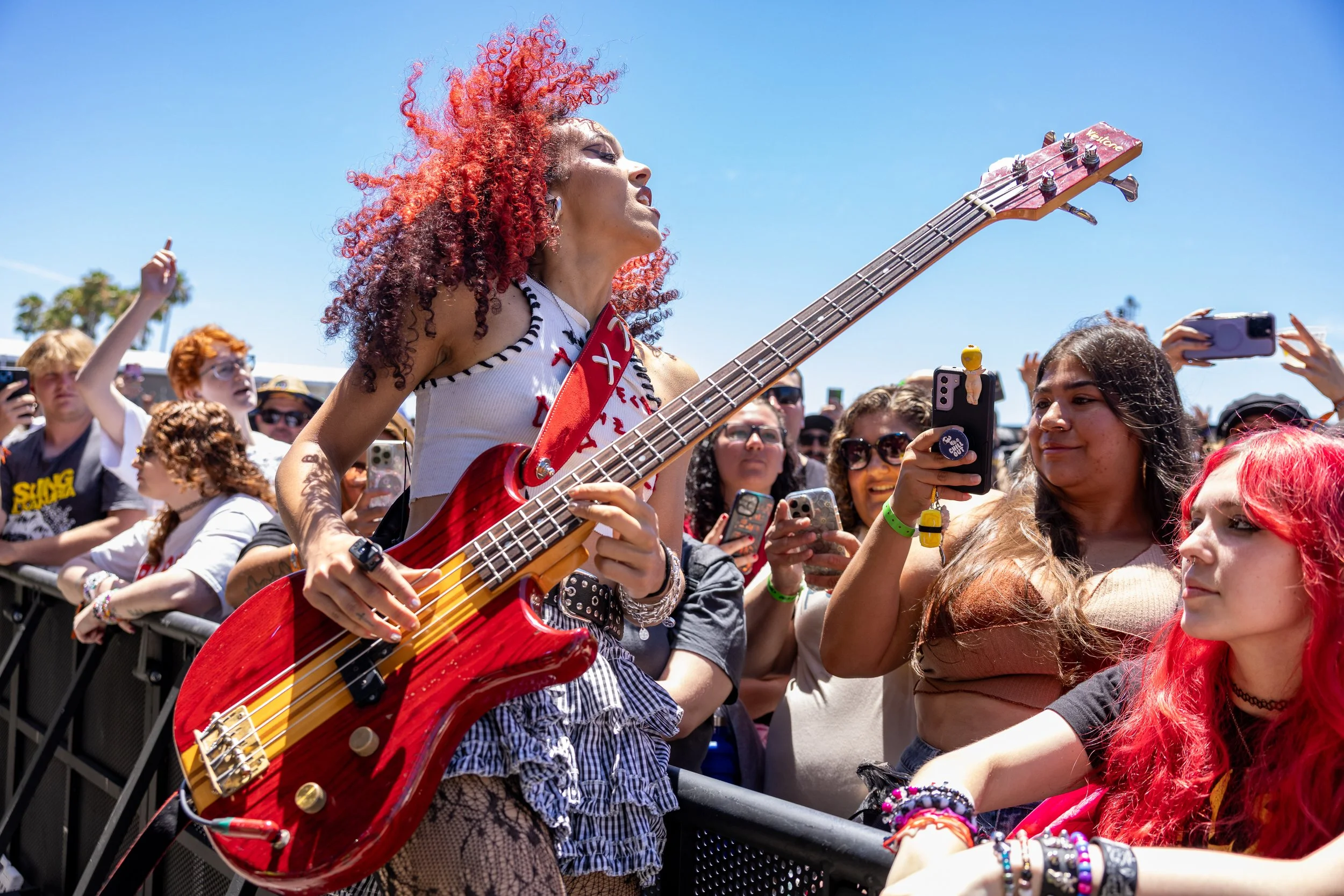 A singer with bright red hair, matching her bright red guitar, leans against the fence with the crowed standing just inches away from her on the other side
