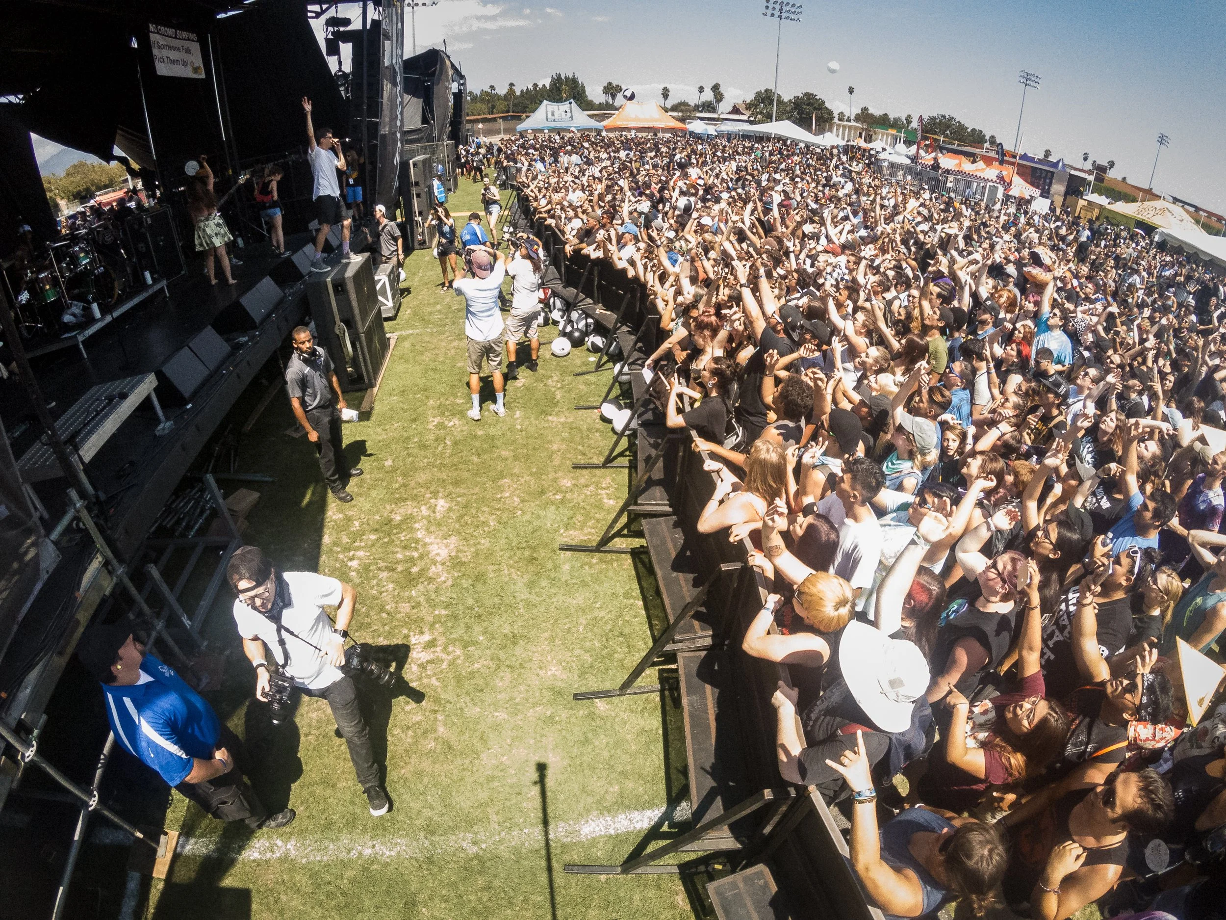 A band is on a dark stage, with a grassy gap between them and a crowd that's in the bright sun