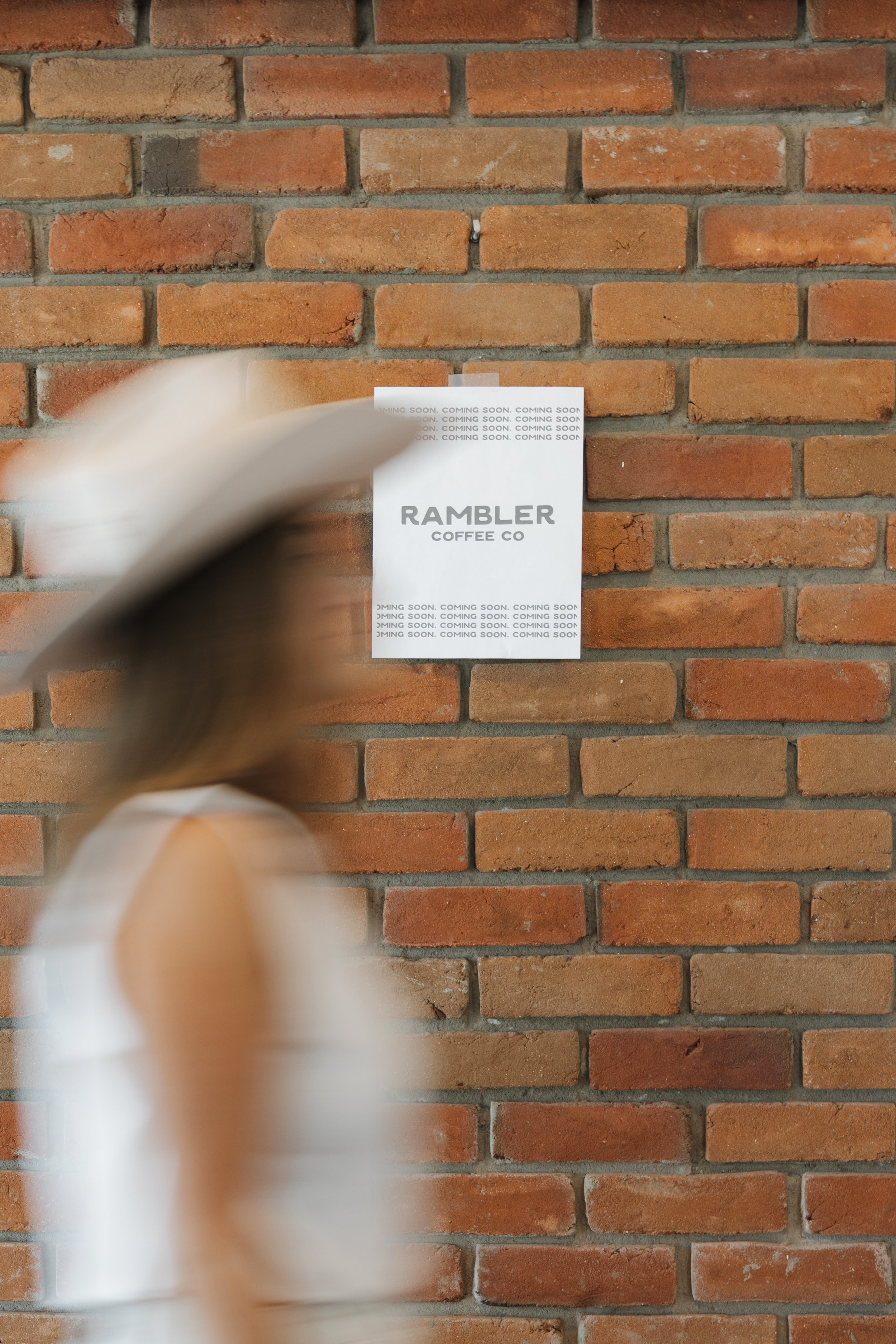 A blurry person walking by a red brick wall with a poster hanging on it reading “Rambler Coffee Co.”