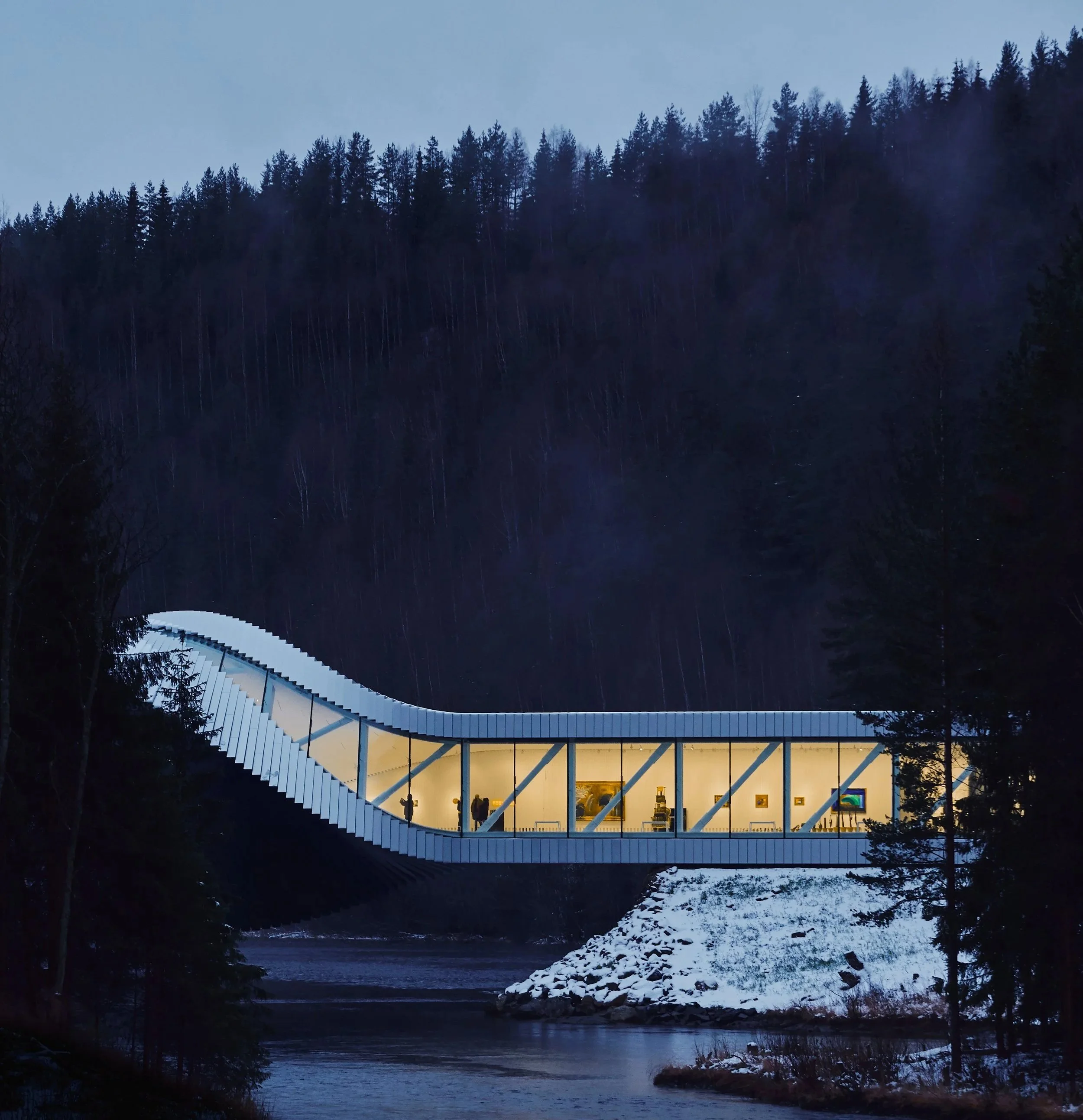 Nighttime view of a modern glass building with interior lighting, built on a slope over a river, surrounded by trees and mountains.