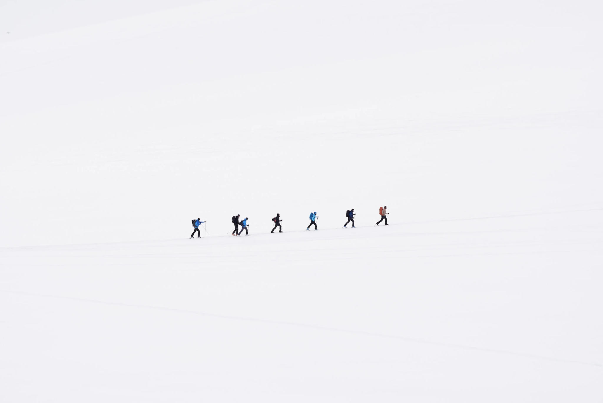 Seven people hiking in snow with backpacks and walking poles, walking in a line across a vast, snow-covered landscape.