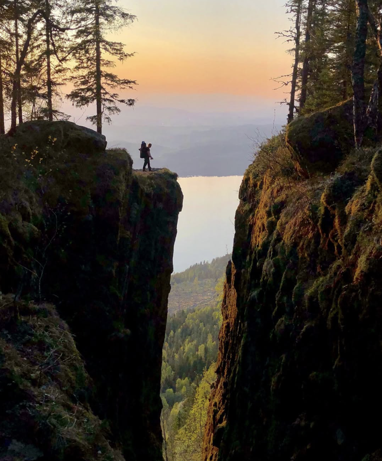 A hiker carrying a child on a hiking trail between canyon walls, overlooking a body of water with a forest and hills in the background during sunset.
