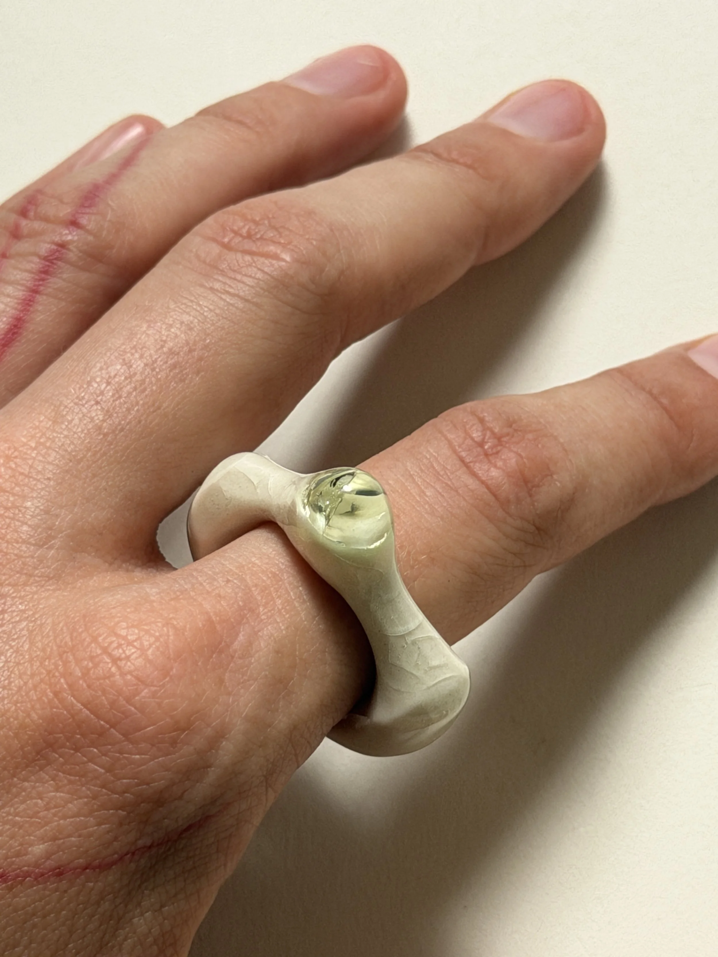 Bone-white porcelain ring with thick band worn on hand, light celadon glass droplet visible
