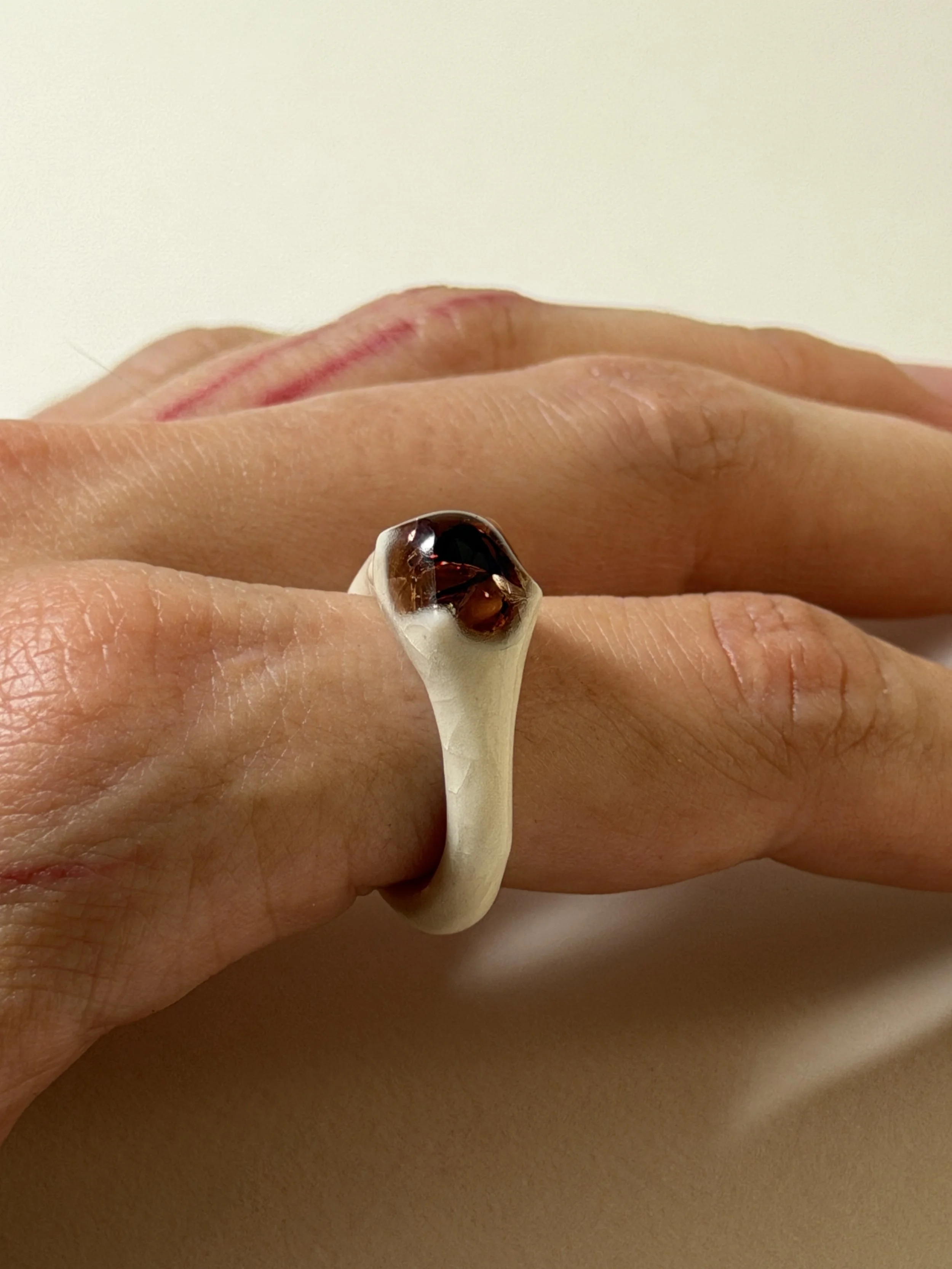 Bone-white porcelain ring with thin band worn on hand, crimson glass droplet visible