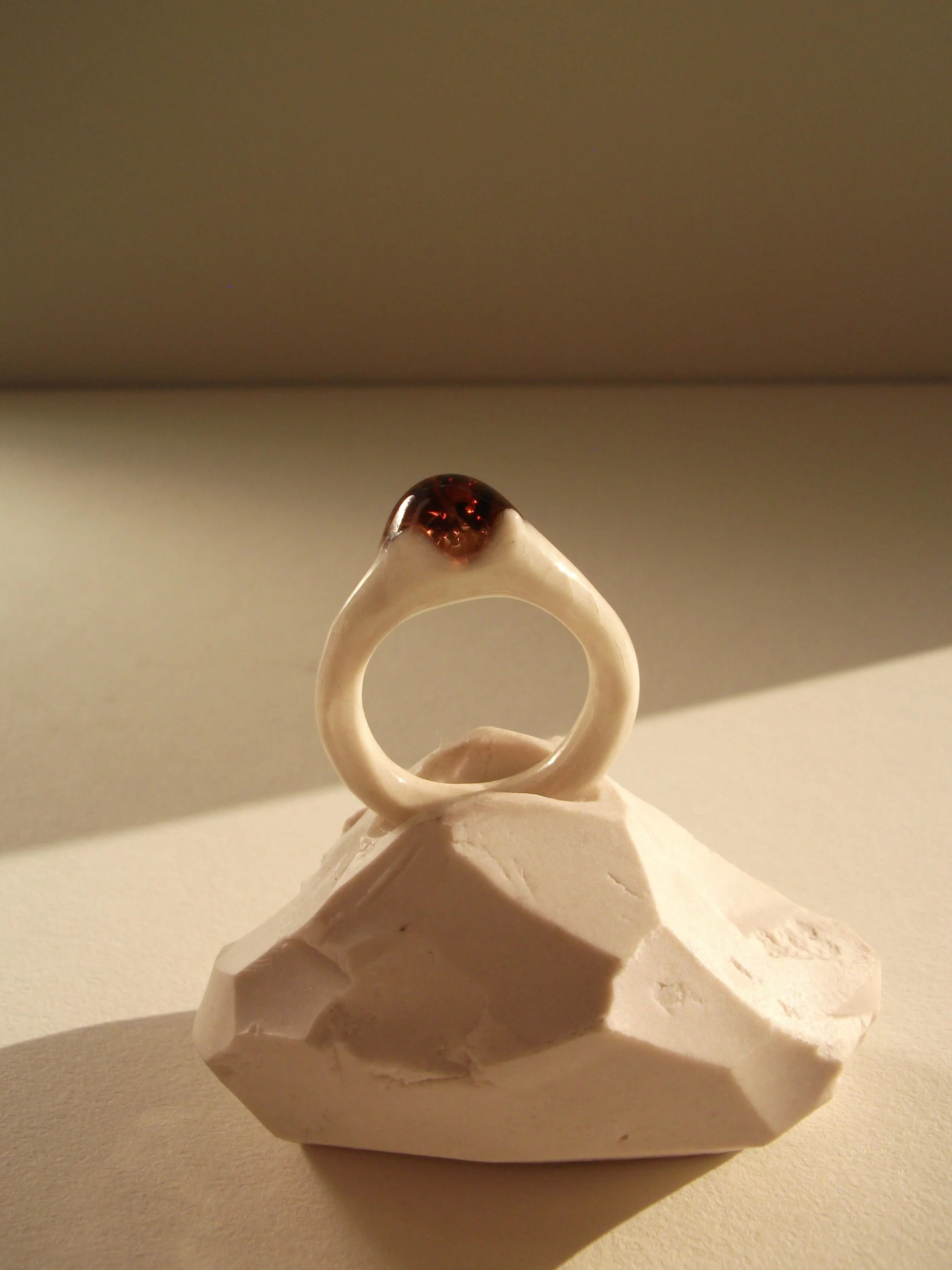 Angled view of thin bone-white porcelain ring with crimson glass droplet at center