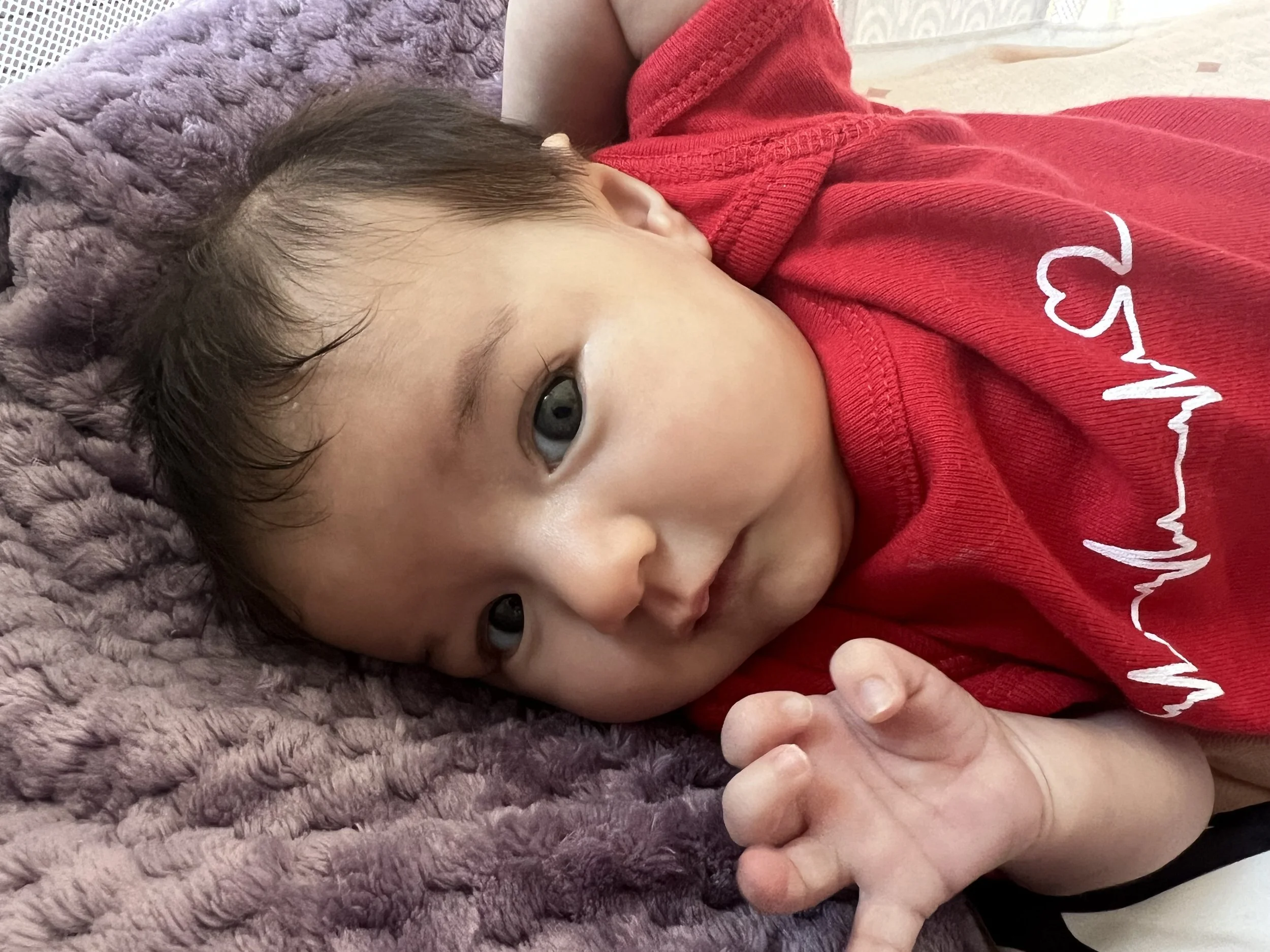 Close-up of a baby with dark hair and blue eyes lying on a fluffy purple blanket, wearing a red shirt with a white heartbeat line and heart design.