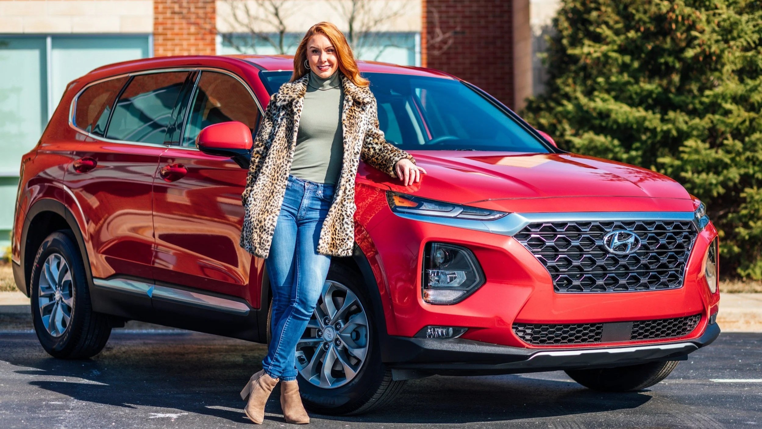 A woman with red hair wearing a leopard print coat, gray top, and jeans, standing next to a red Hyundai SUV in a parking lot.