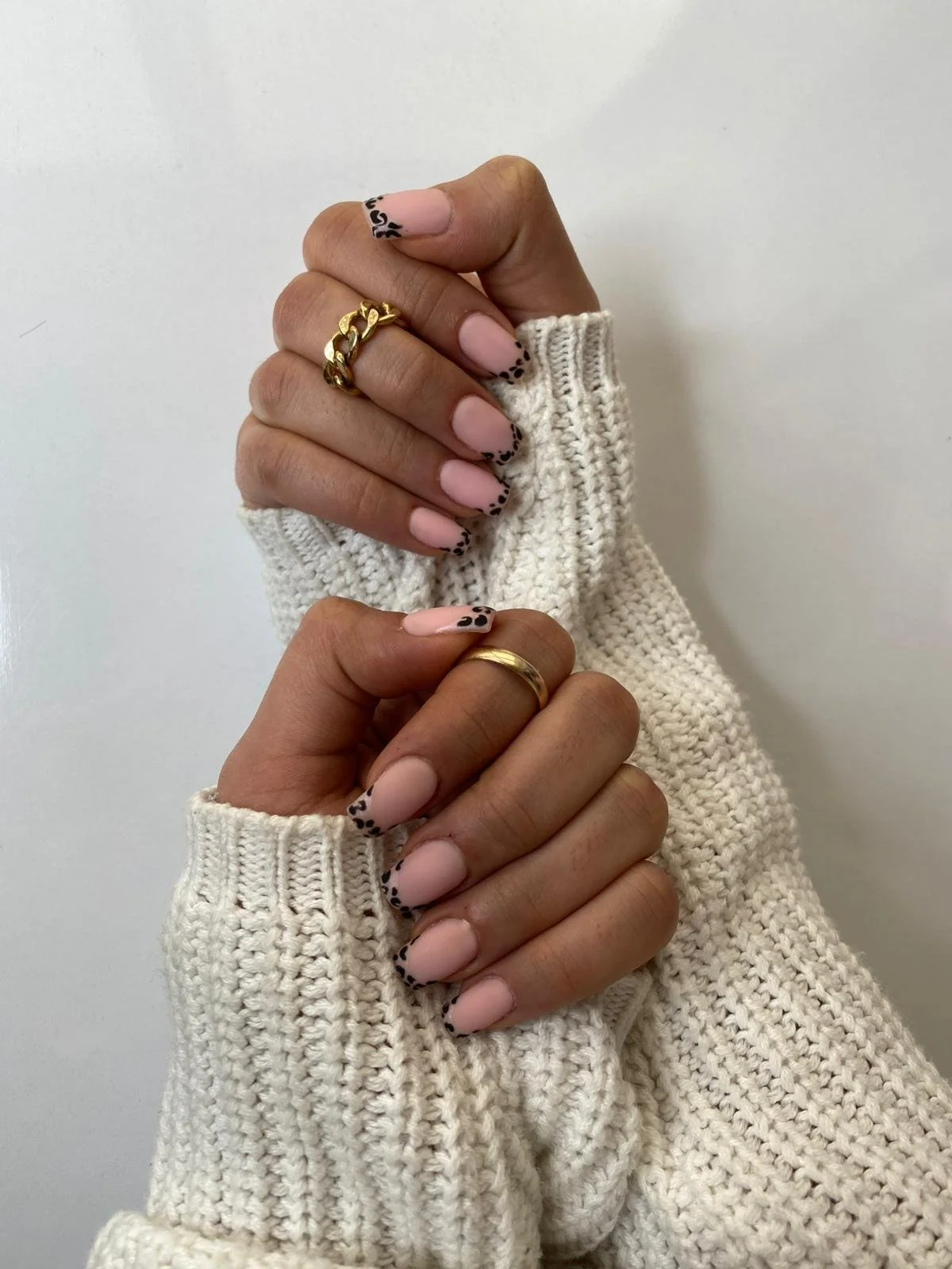 Two hands with manicured nails featuring black leopard print tips, adorned with rings, and dressed in cream knit sweaters, posed against a plain white background.