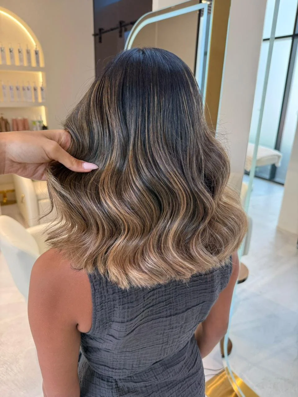 Woman with shoulder-length wavy hair, showing a hair color transition from dark roots to blonde tips, in a salon setting.