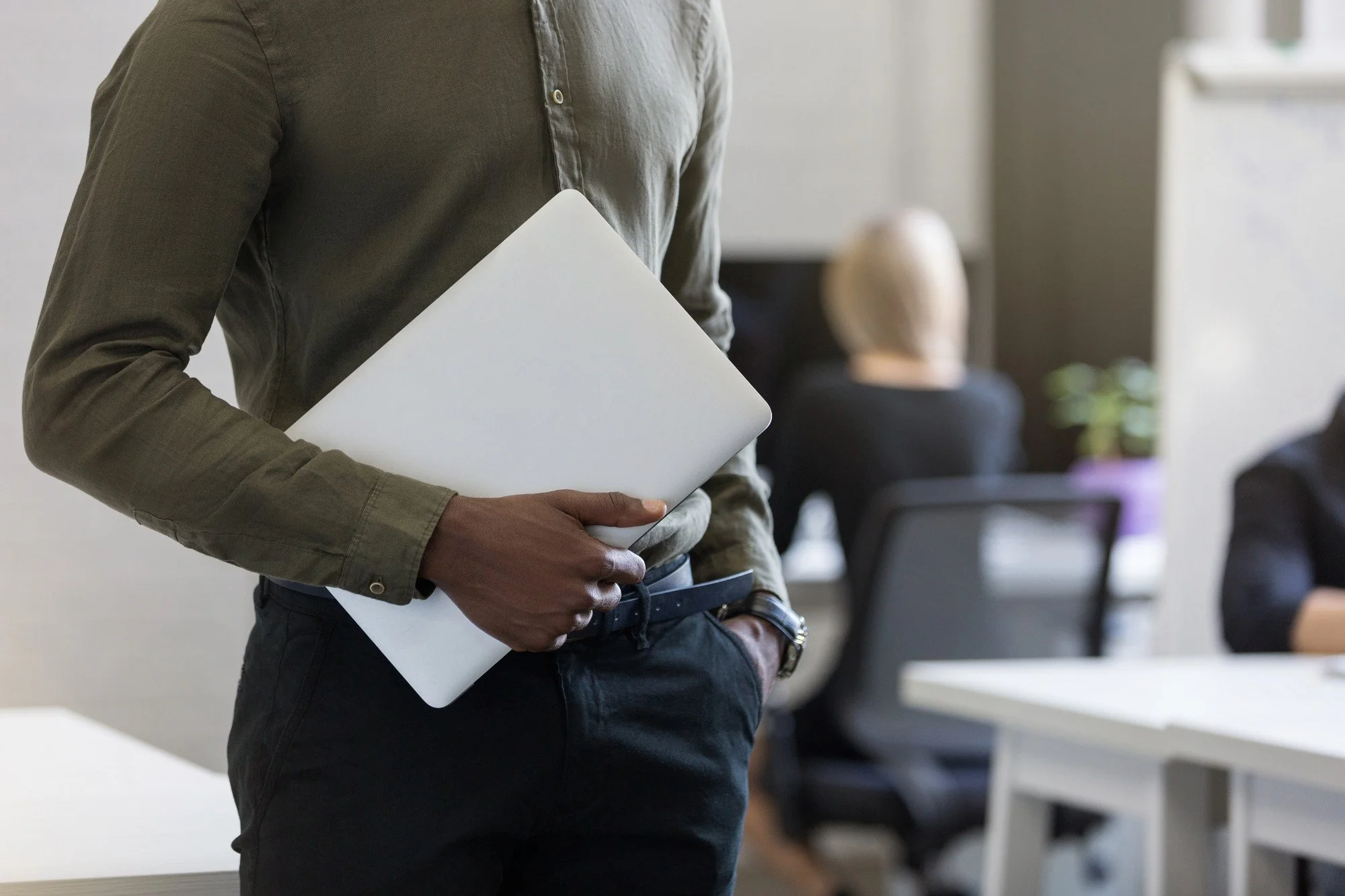 Person holding a closed laptop in an office setting with other people working at desks in the background.